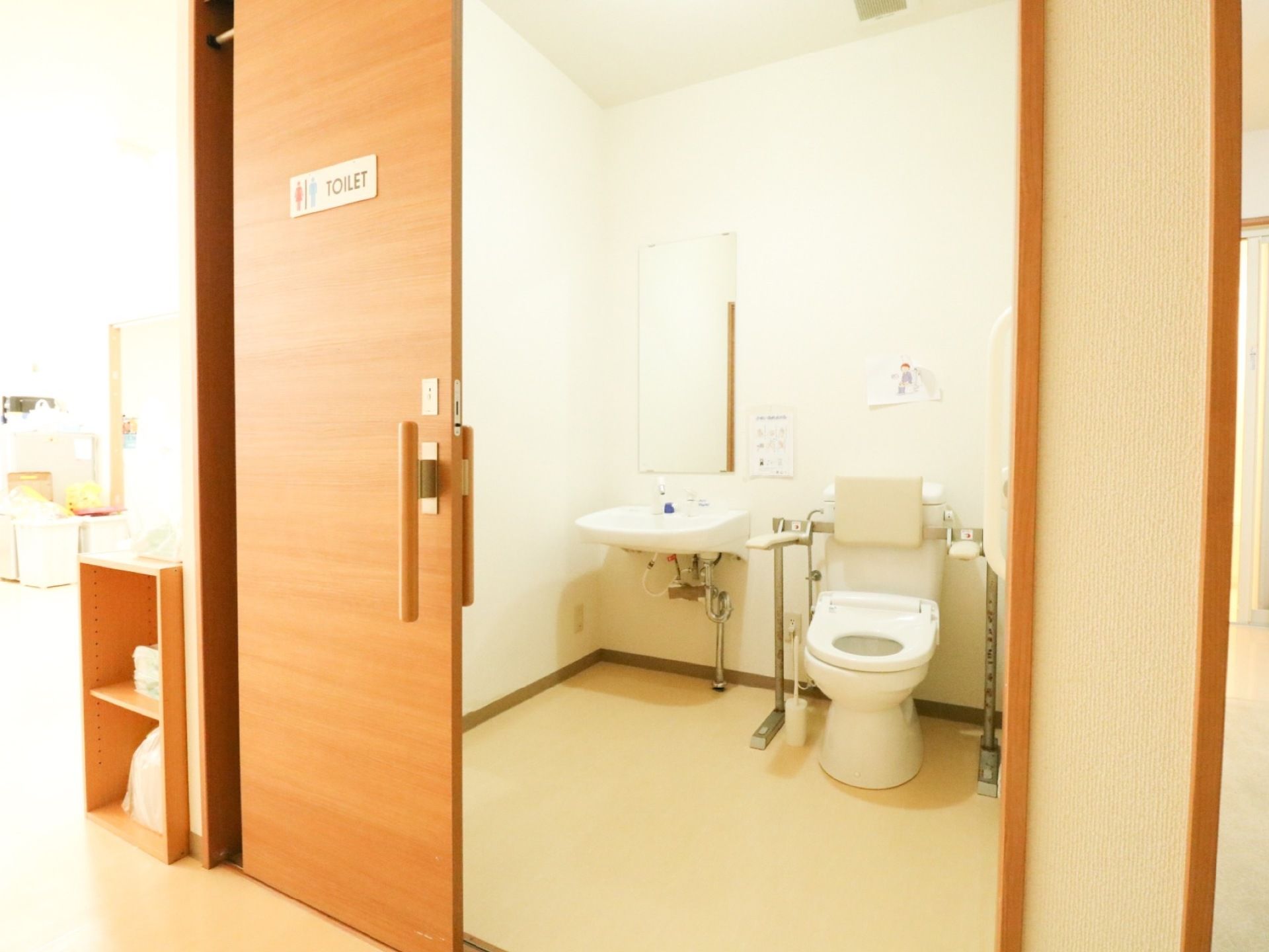 木製の引き戸で仕切られた広い空間に、洋式便器と手洗器が設置されている。背もたれとアームレストが便器に取り付けてある。