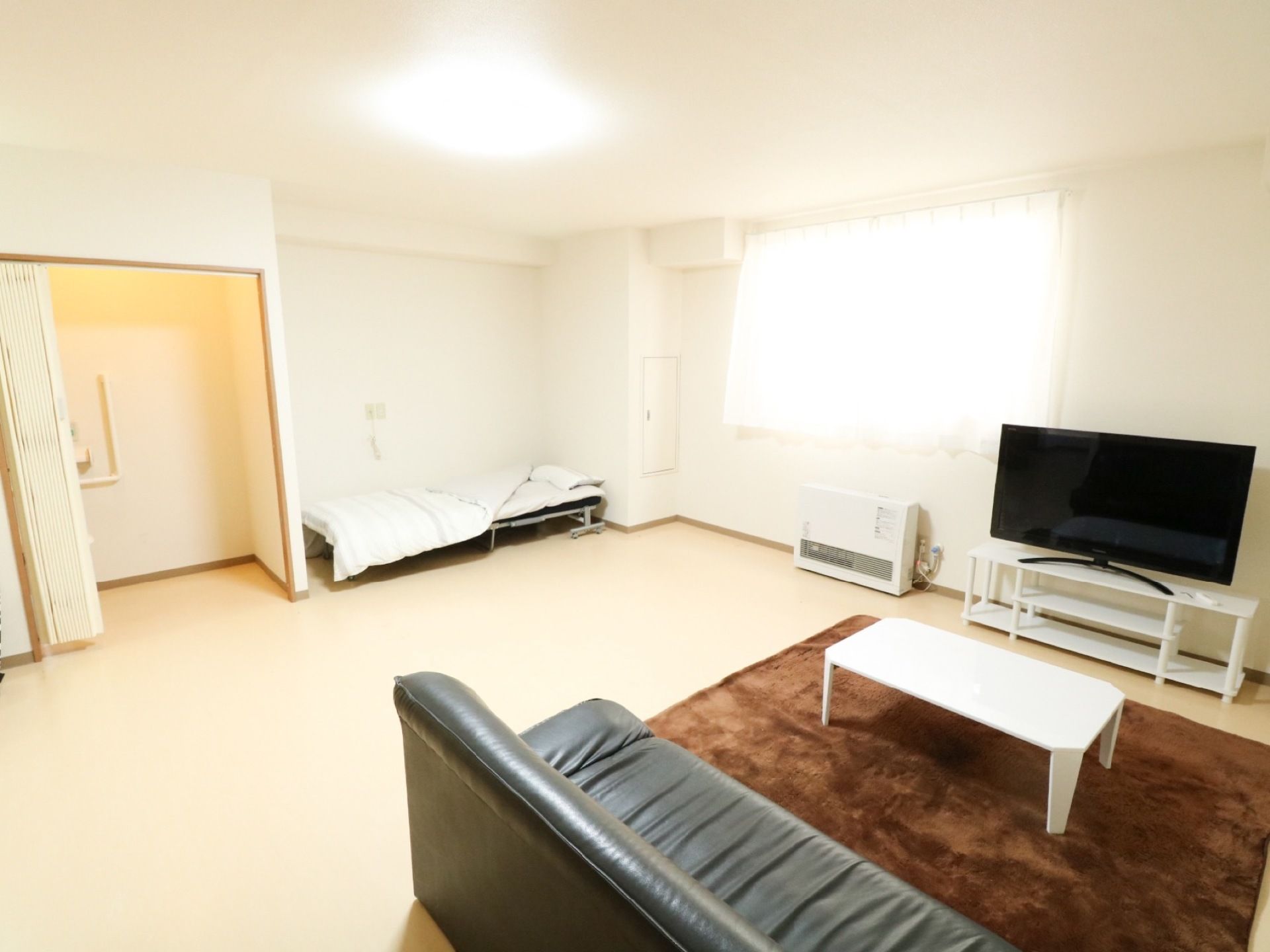 広い室内に、ソファや液晶テレビ、ベッドが設置されている。アコーディオンタイプの間仕切りの奧にＬ字型の手すりが見える。
