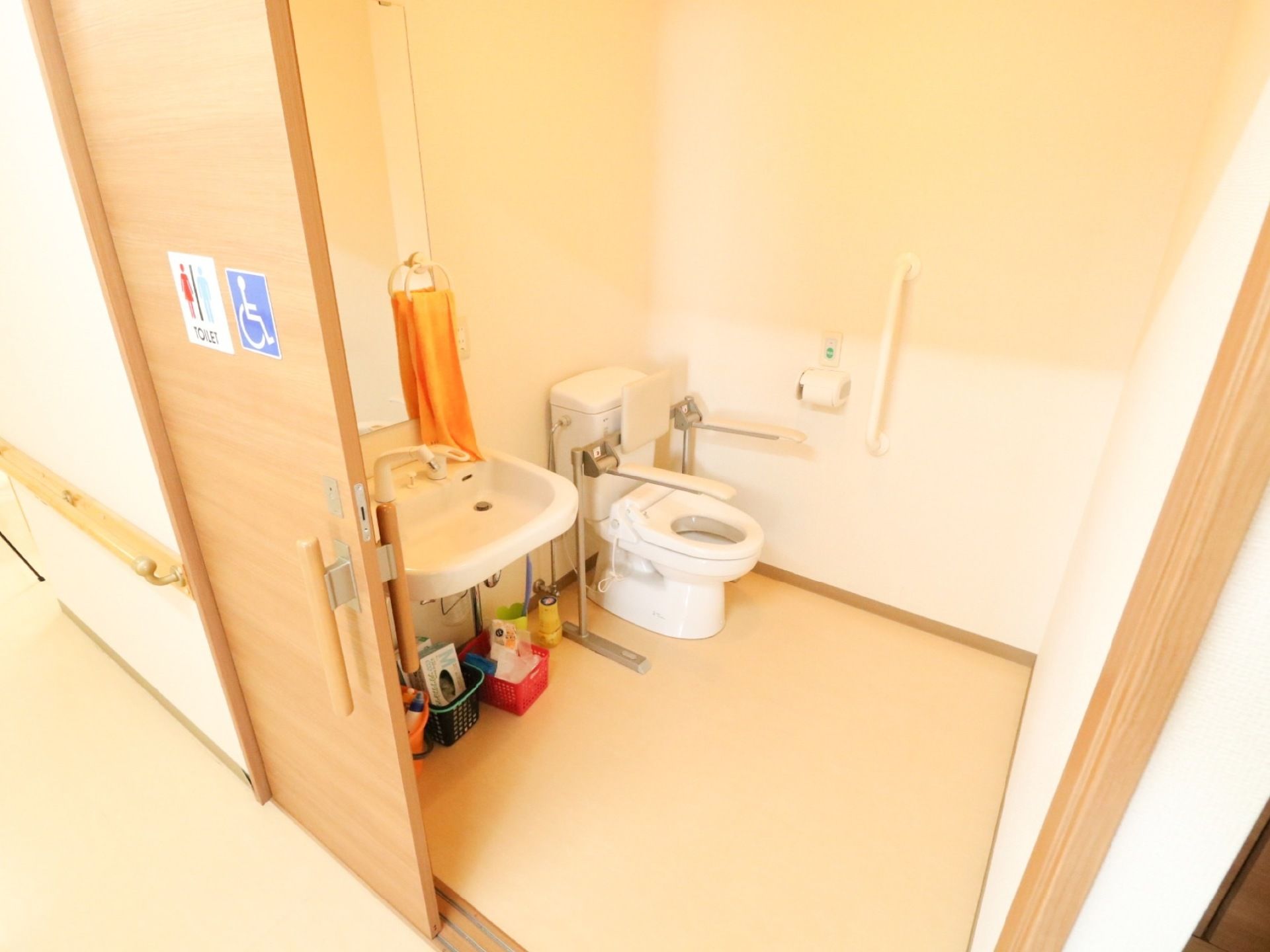 木製の引き戸にトイレと車椅子の表示が貼り付けてある。広い空間に洋式便器と手洗器が並べて設置されている。