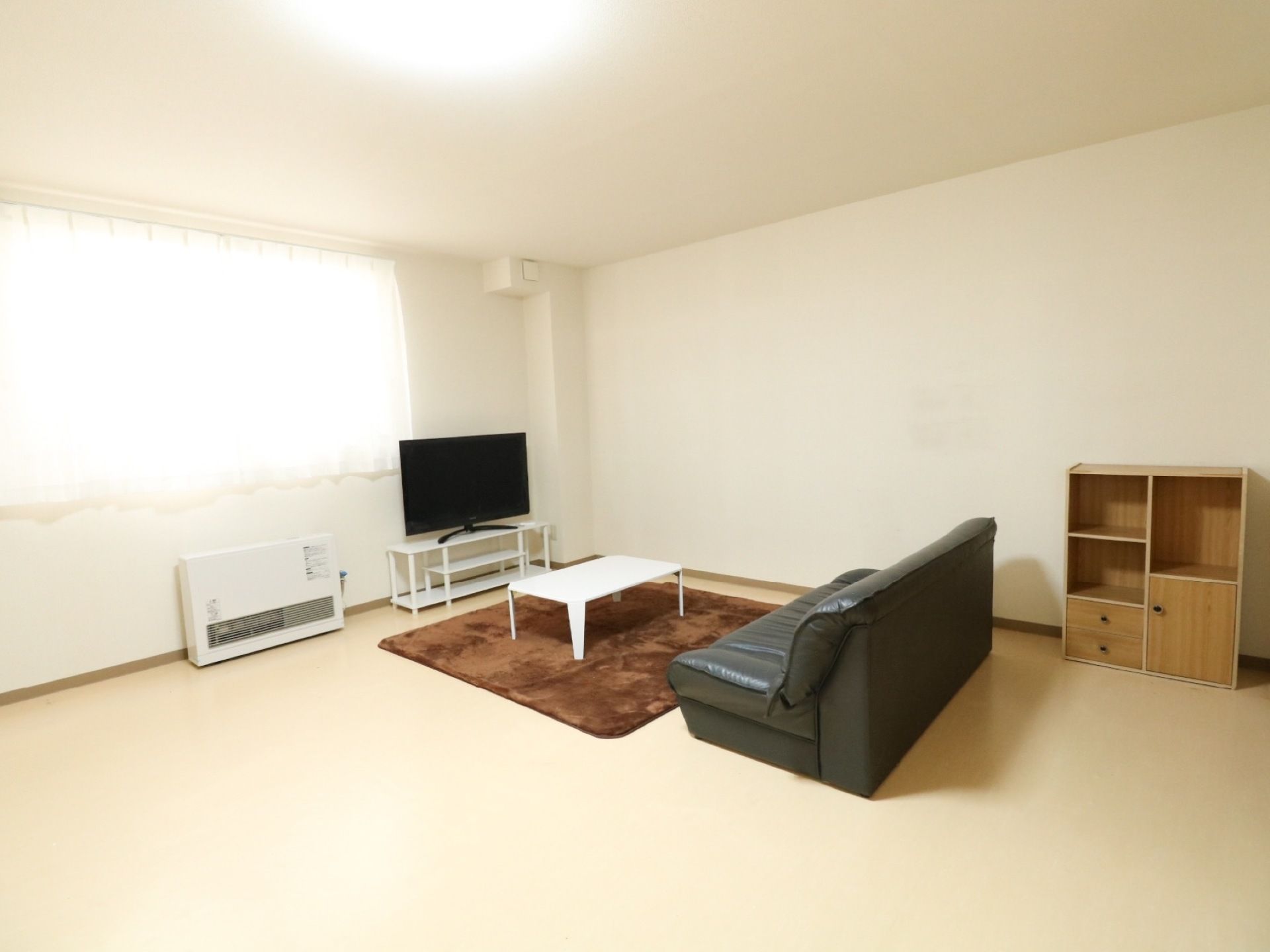 窓がある広々とした室内に、革張りのソファやテーブル、液晶テレビが設置されている。右側に木製の収納棚が置いてある。