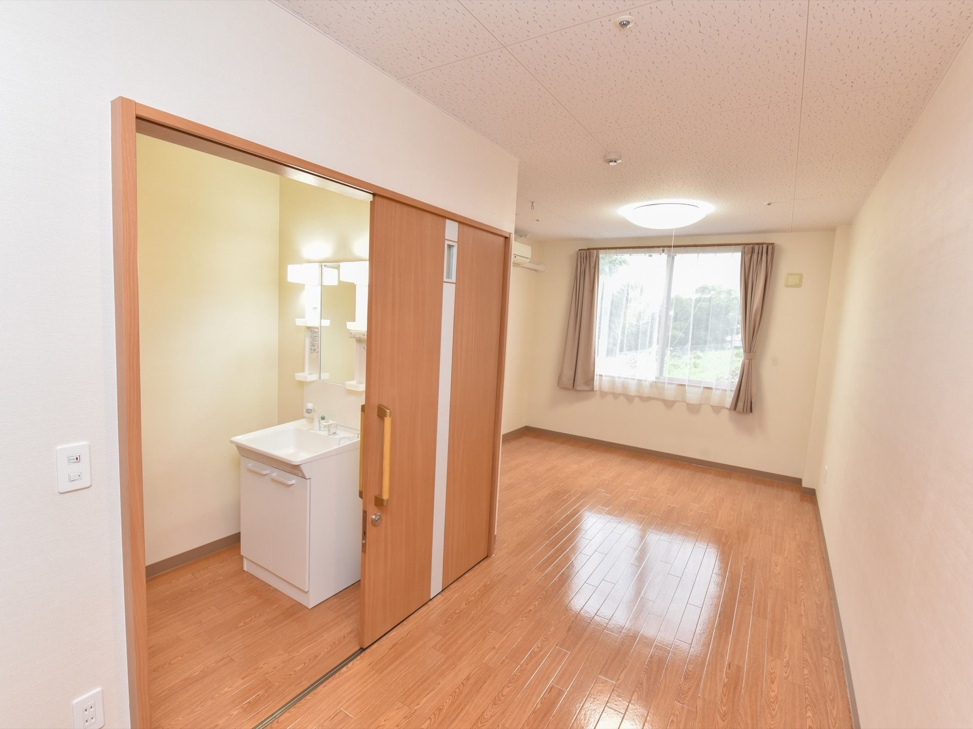 広々とした居室には窓がある。窓にはカーテンが掛かっている。洗面台もあり、鏡や収納棚が確保されている。