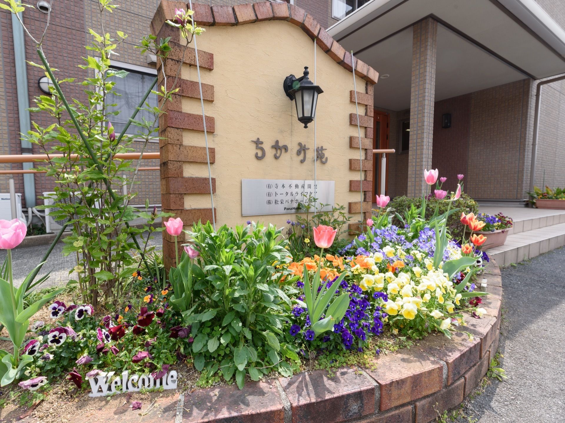玄関前に、レンガで囲われた花壇が整備されている。施設名の書かれた大きな表札があり、ランプが付いている。