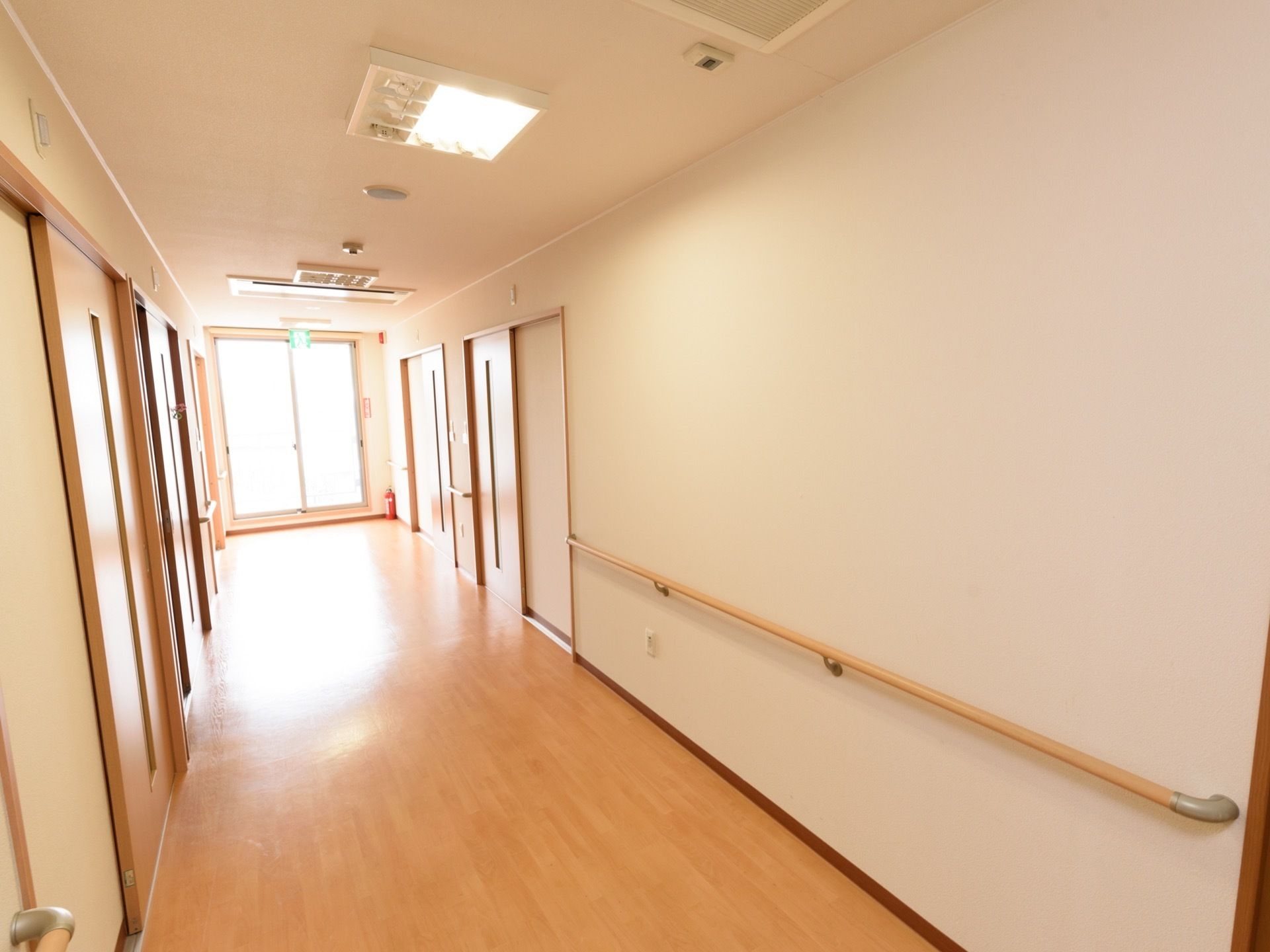 フローリングの床に白い壁の廊下である。両脇の壁には手すりと、居室の入り口がある。入り口はスライドドアである。
