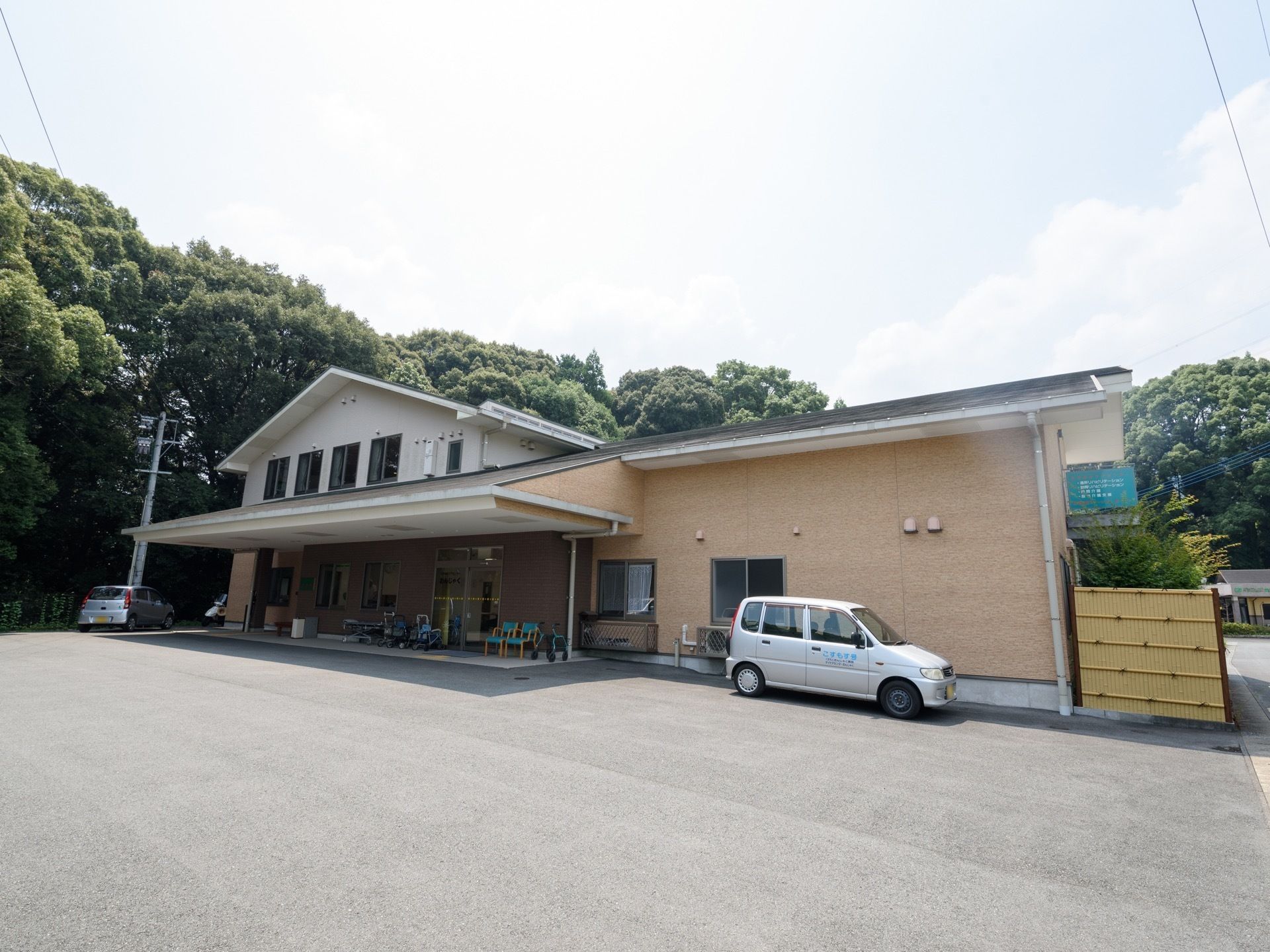 自然に囲まれた2階建ての施設である。入り口前には大きな屋根が取りつけられている。駐車スペースも確保されている。