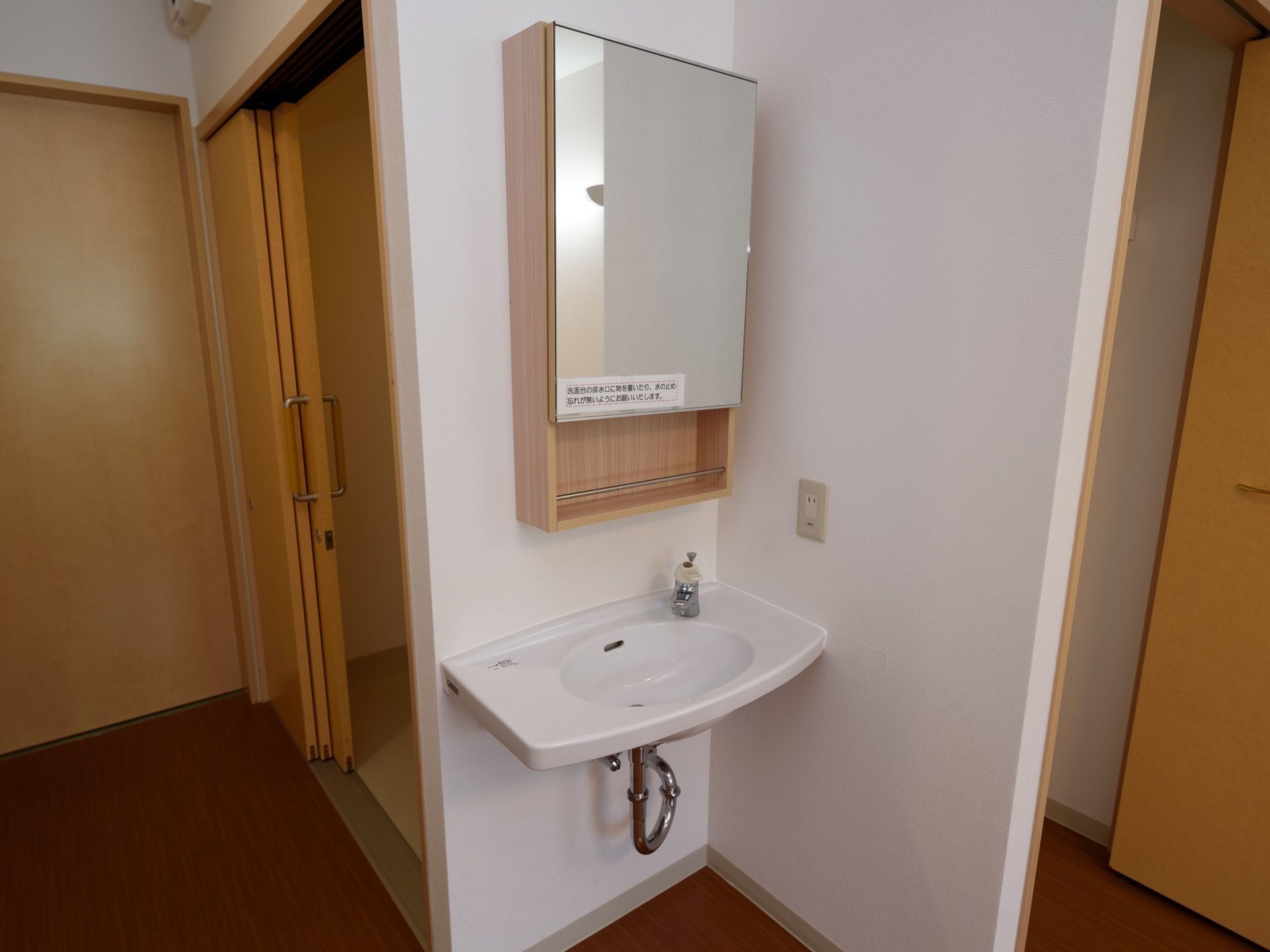 床がフローリングの居室には、専用の洗面所が配置されている。洗面台の上には整理棚と鏡がある。奥のトイレのドアは引き戸式である。