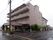 正面に建物が建っており、左側には道路が通っている。建物左側の２階以上の部分にはベランダがある。右側には木々が植えられている。
