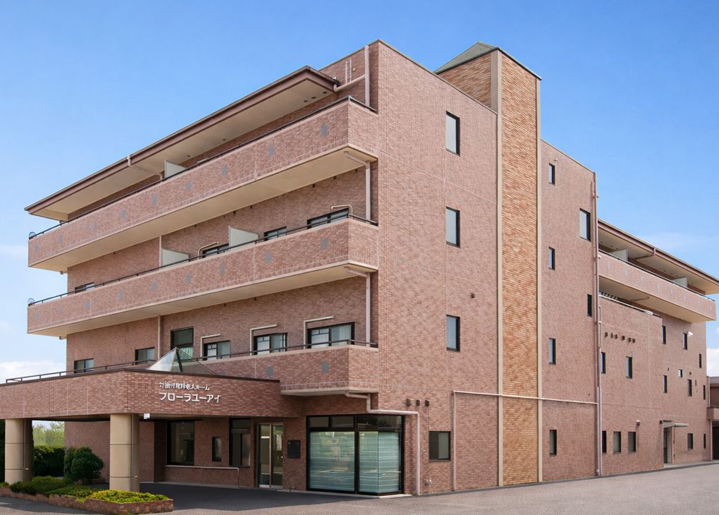 正面に建物が建っており、左側には道路が通っている。建物左側の２階以上の部分にはベランダがある。右側には木々が植えられている。