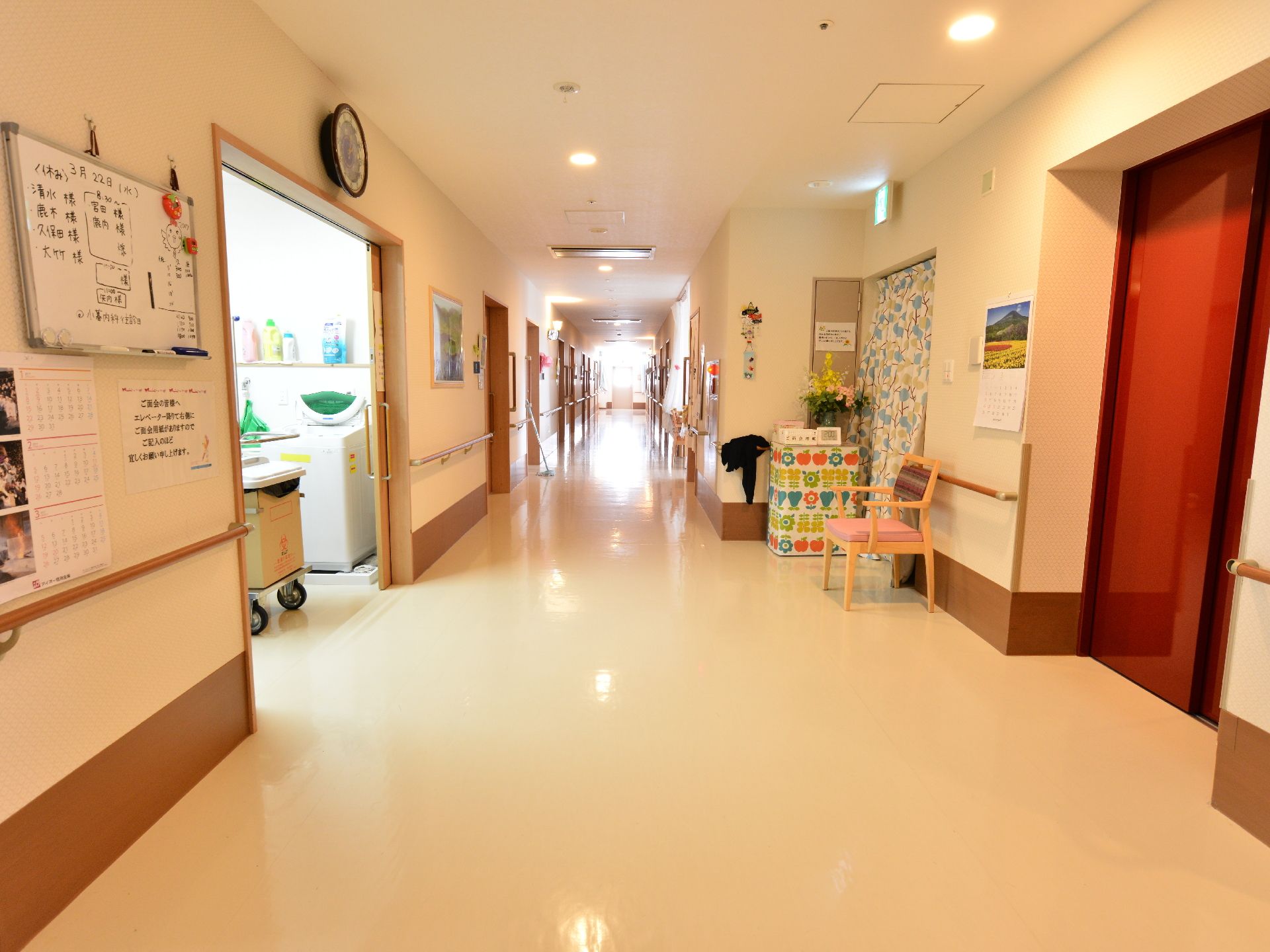 個室の扉が並ぶ廊下。手前にはエレベーターの扉があり、その隣には椅子が一脚置かれている。壁には絵が飾られている。