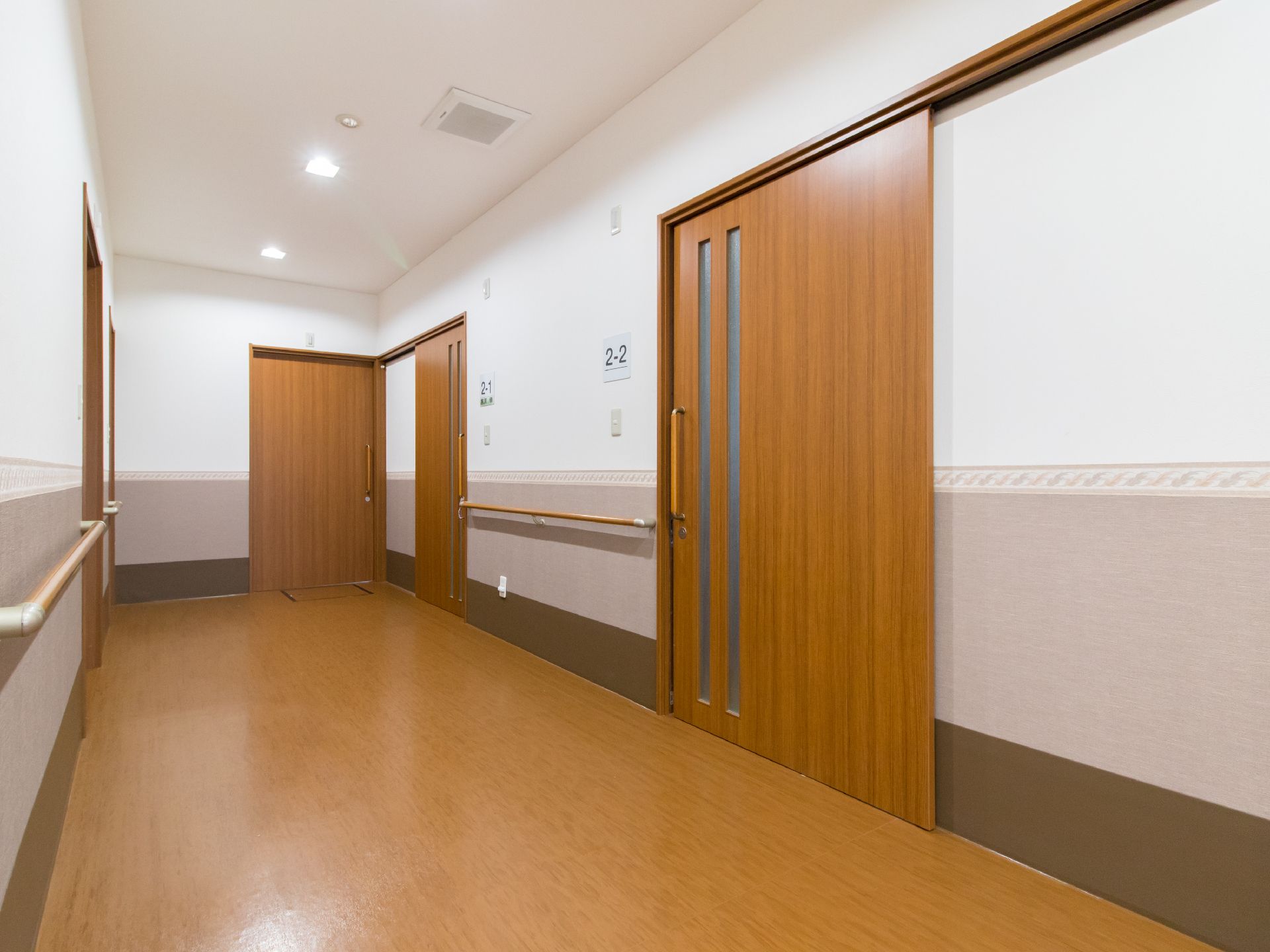 廊下の突き当りには茶色い扉が並んでいる。2-2と書かれた部屋と別の部屋の間には手すりが取り付けられている。