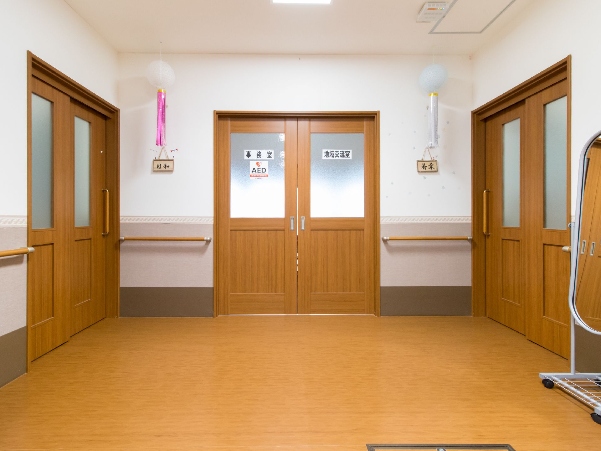 キャスター付きの鏡が置かれた近くには事務室、地域交流室と書かれた扉がある。AED装置設置の表示もある。