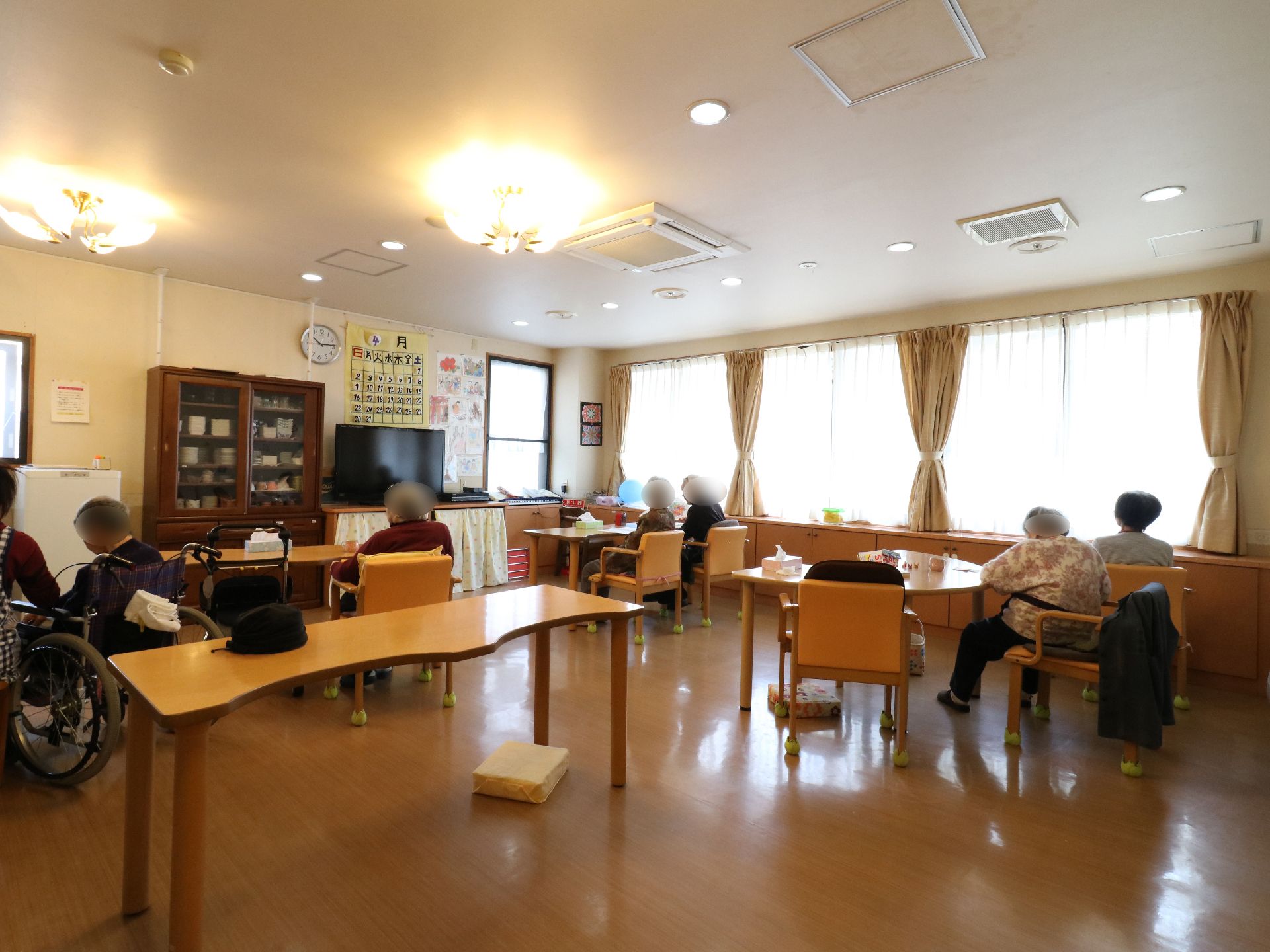 食堂でダイニングテーブルと肘付き椅子が置いてある。所々車椅子で席についているところもある。カップボードの隣にテレビが置いてある。