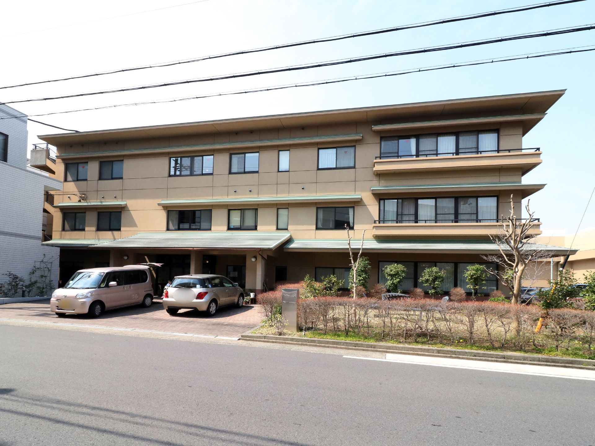 駐車場が設けてある。施設の外壁は、茶色で、緑色の屋根である。歩道の一部に花壇があり、木が定間隔で植えてある。