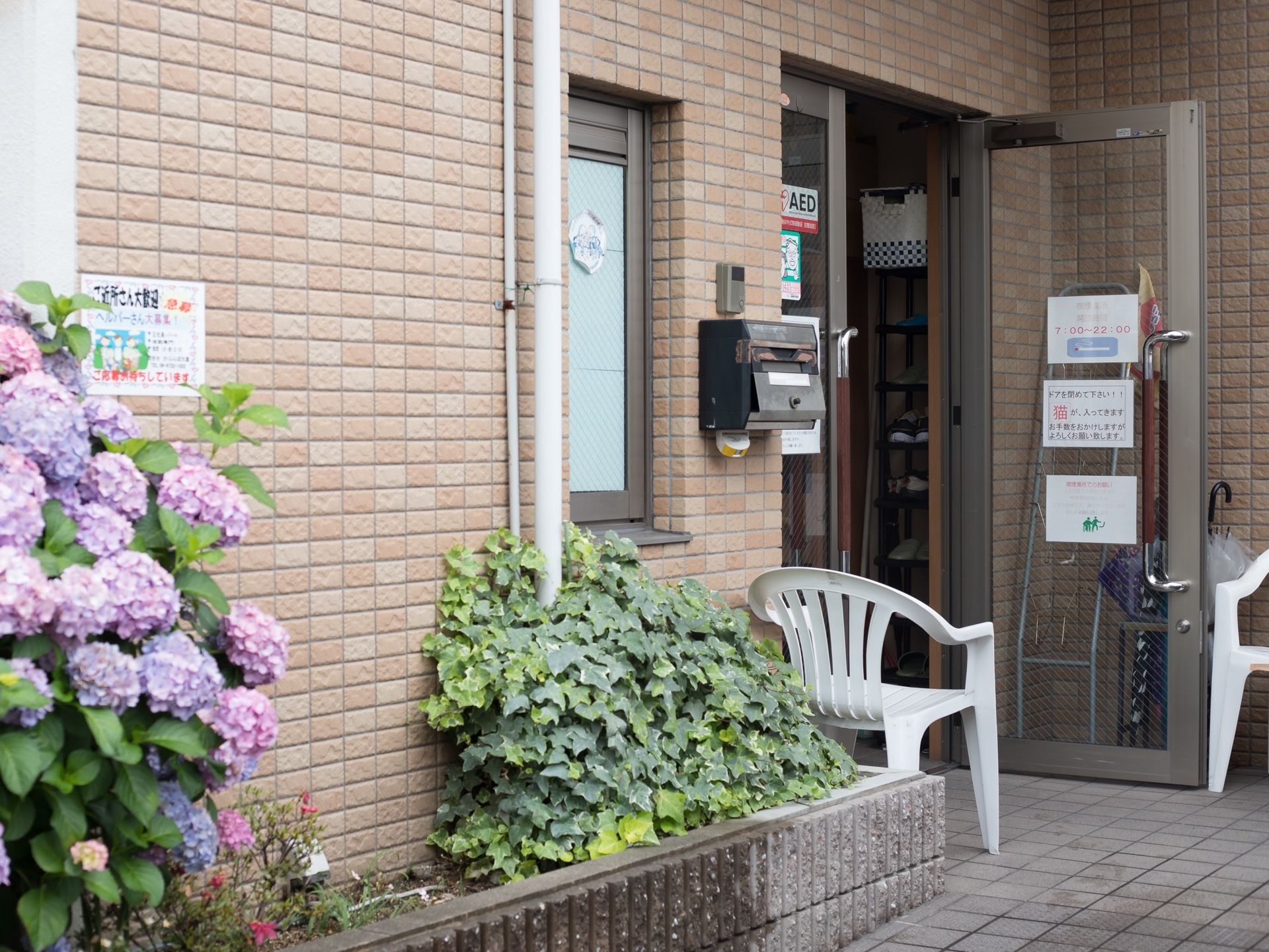 玄関前には椅子が２脚置かれている。マガジンラックと傘立てが用意されている。あじさいなどの植栽が植えられている。