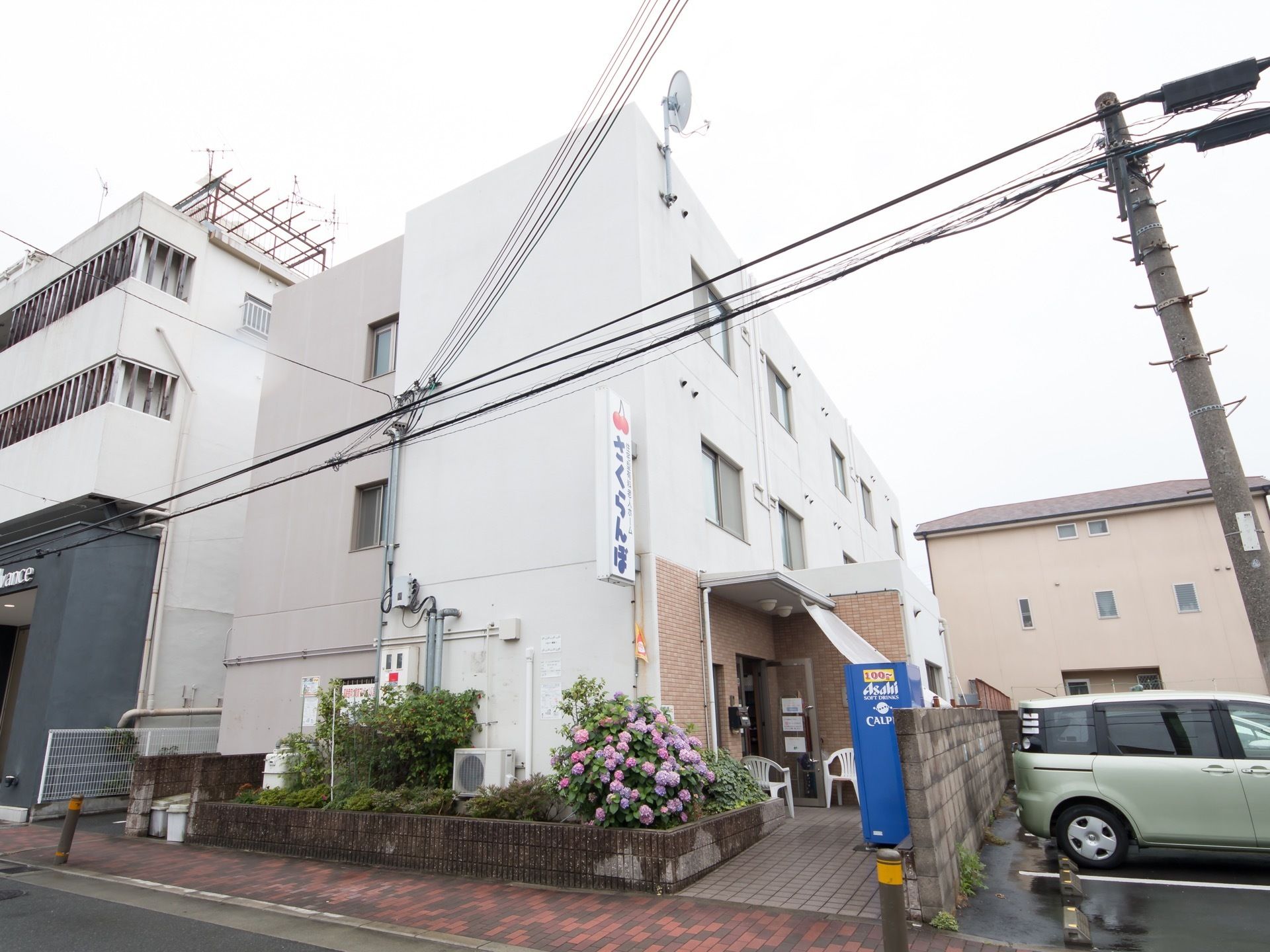 ３階建ての施設で、外壁は白く塗られている。２階部分に大きな看板が取り付けられている。玄関前に自動販売機がある。