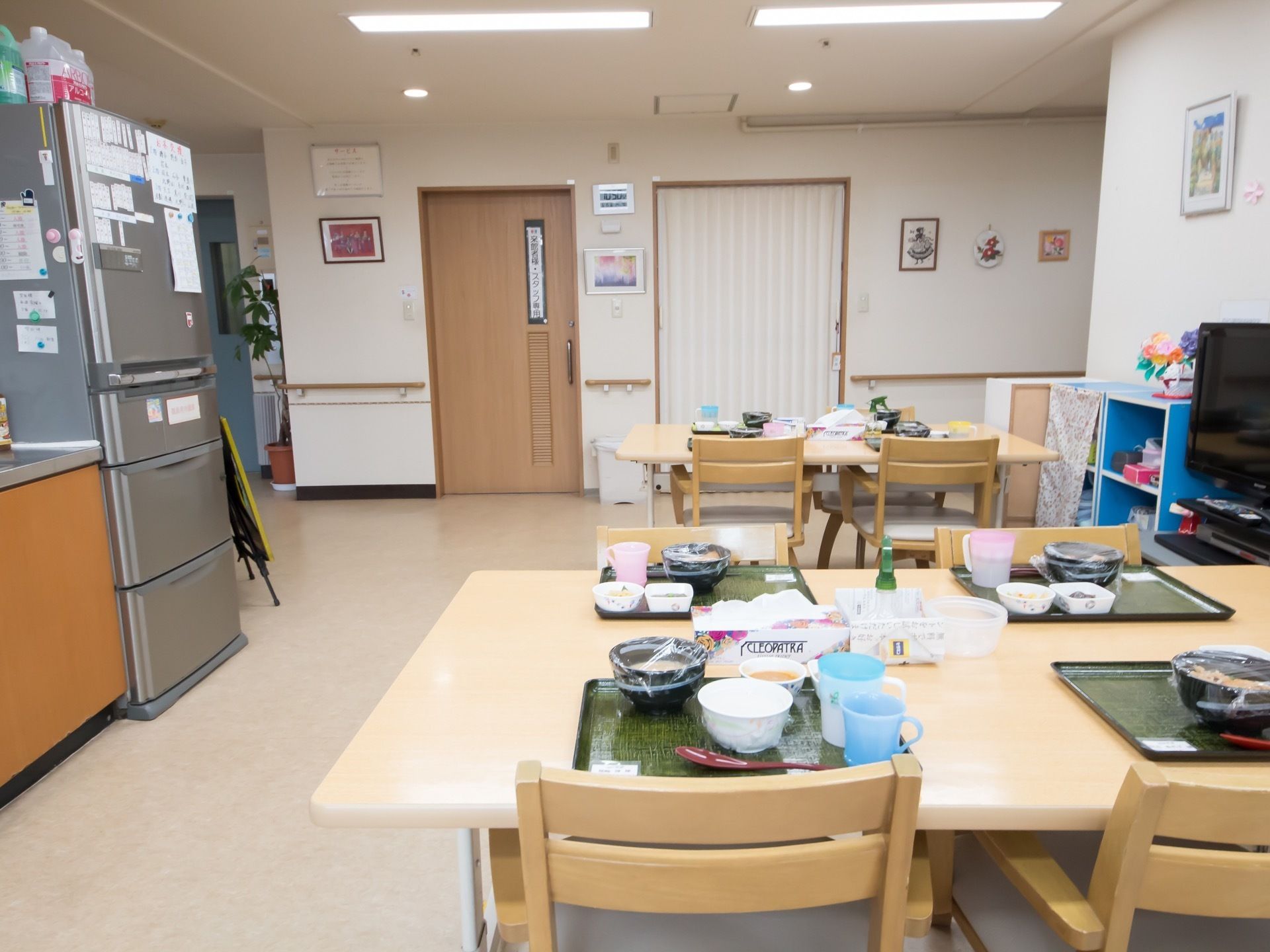 共有スペースにはテーブルセットが並び、食事がトレーに置かれている。家族用の大型冷蔵庫と、壁際に液晶テレビがある。