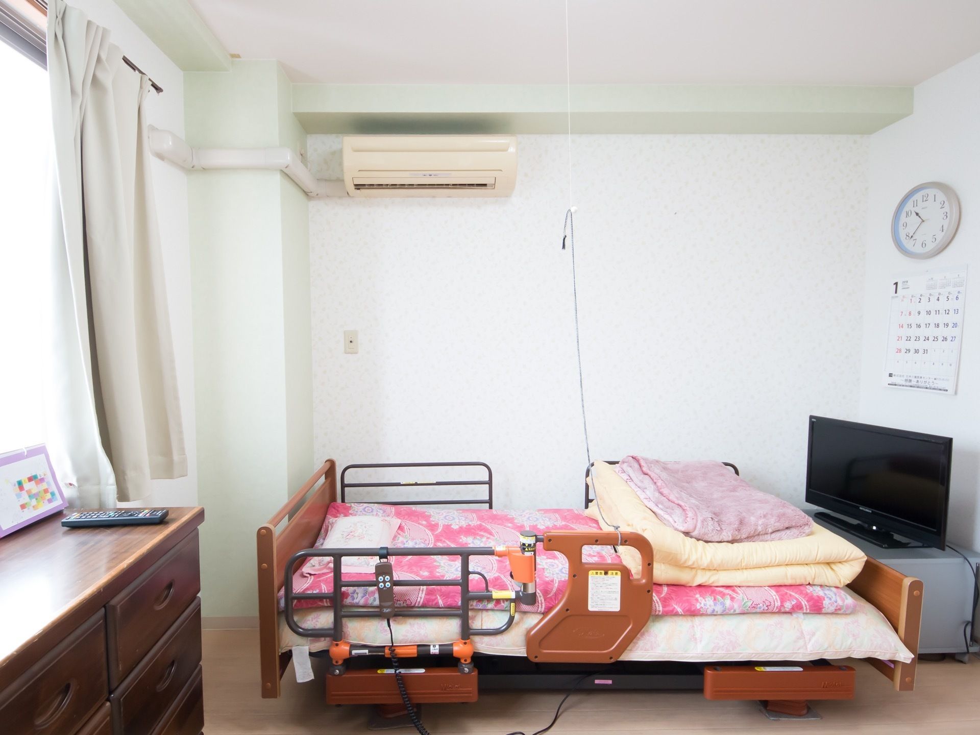 居室には介護用ベッドが用意されている。落下防止の手すりや、ナースコールが取り付けられている。照明の紐もベッドまで下げられている。