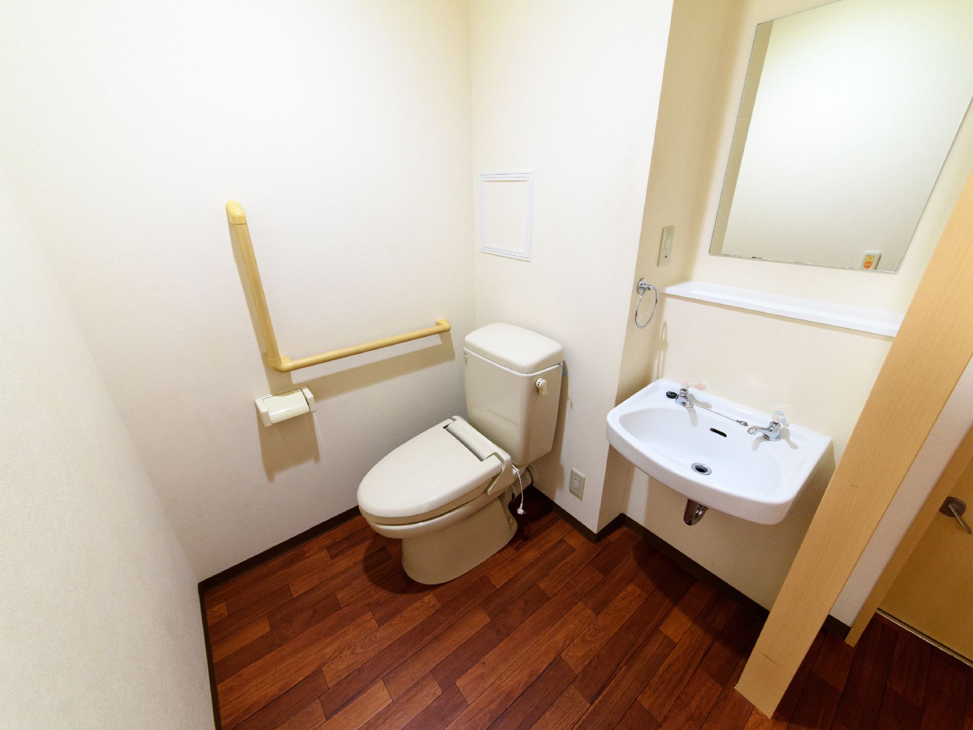 トイレ個室内に、洋式トイレと手洗い場がある。便器横に、L字の手すりがついている。洗面台には鏡と備品置き場がある。