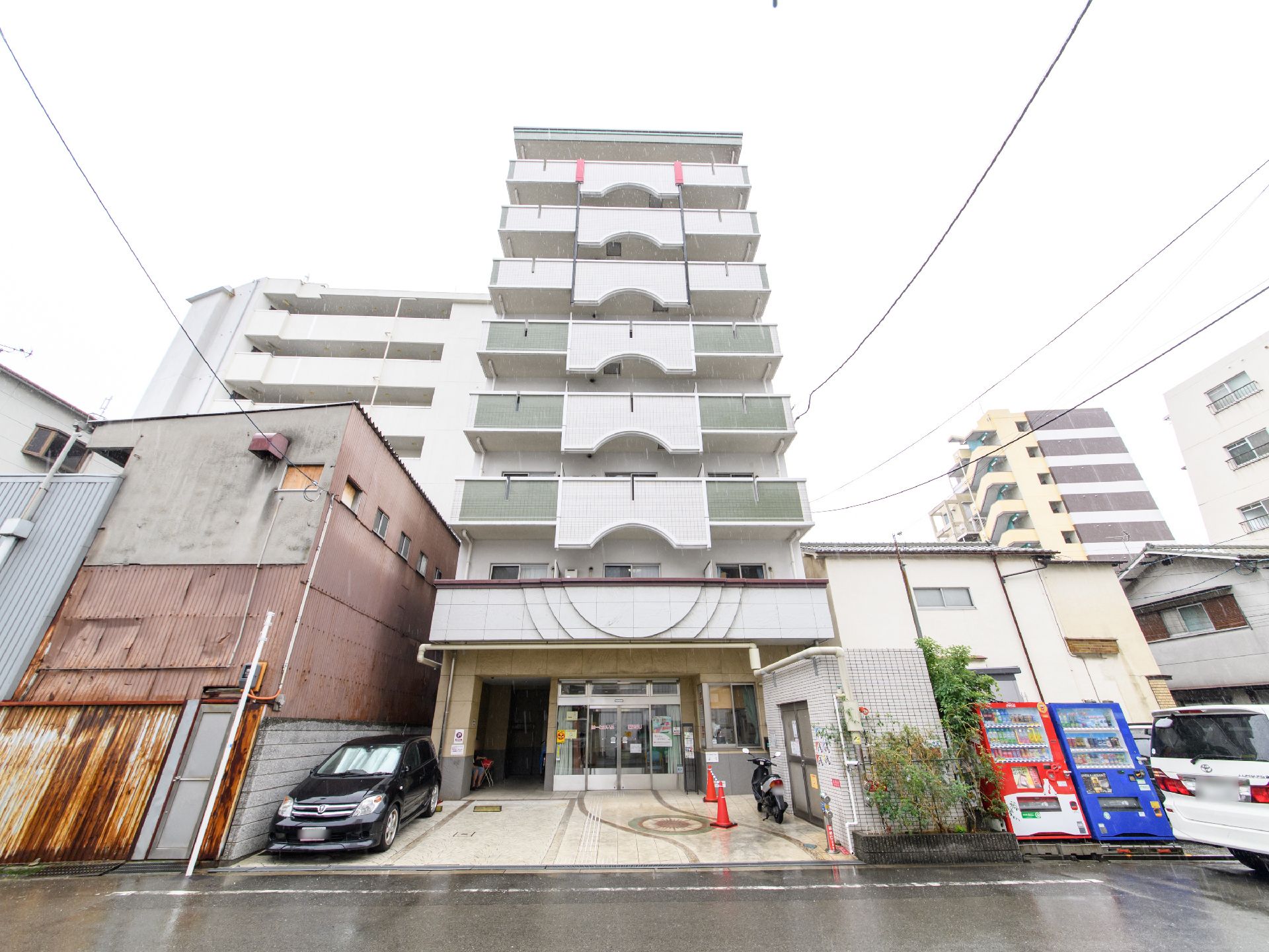 施設は8階建てで、正面から見ると縦長の建物である。外壁は白色で、周囲にも家屋がある。隣に自動販売機が2台置かれている。