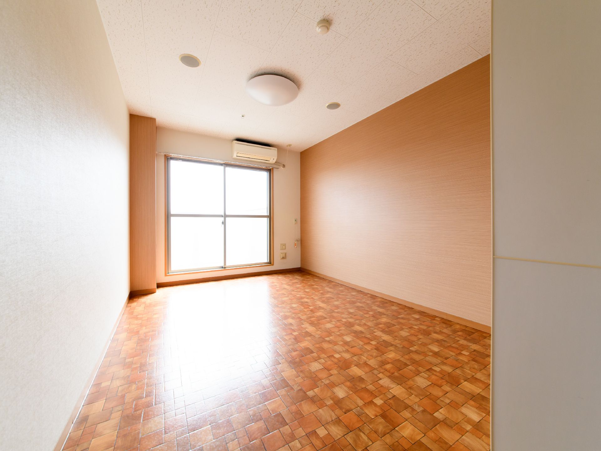 入口から見た、居室の様子。奥に大きな2枚のガラス扉があり、日が差し込んでいる。扉の上に、エアコンが設置されている。