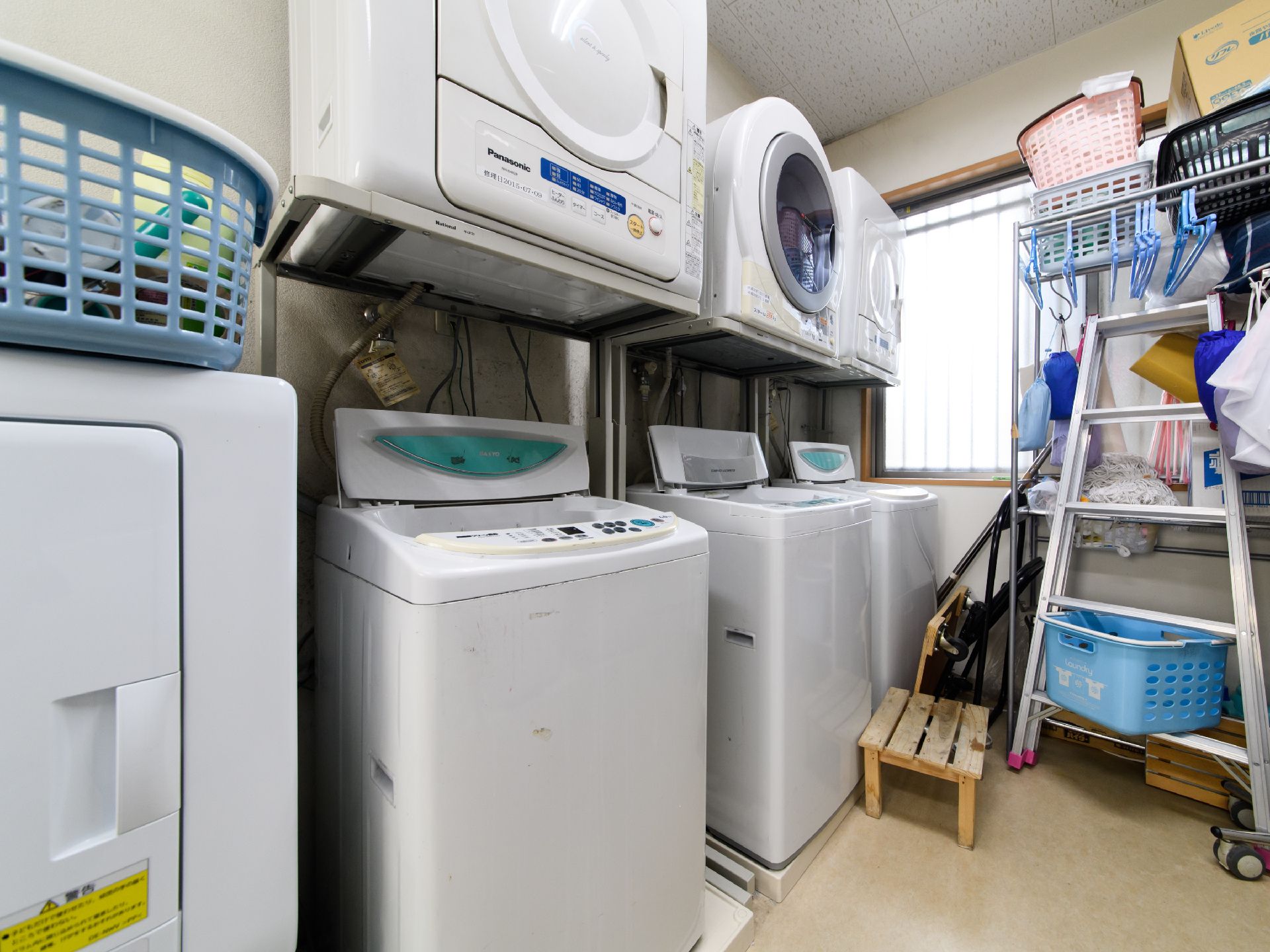乾燥機付きの全自動洗濯機が3台並んでいる。右側には窓と高い棚があり、ハンガーがたくさんかけられている。