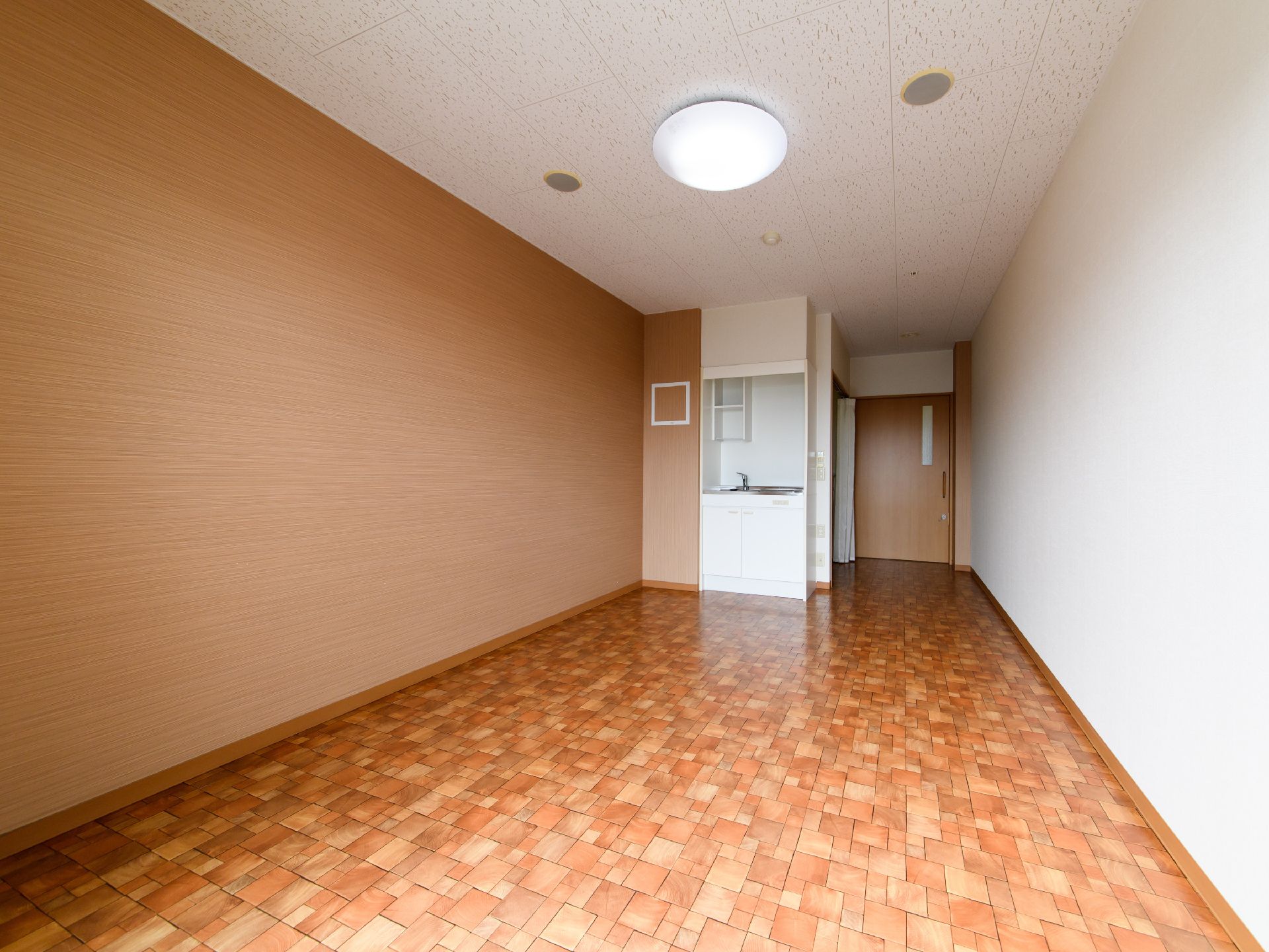 居室の入口は、横開きの木製扉である。入口のすぐ近くに、カーテンが見える。天井には、丸型のライトが取り付けられている。