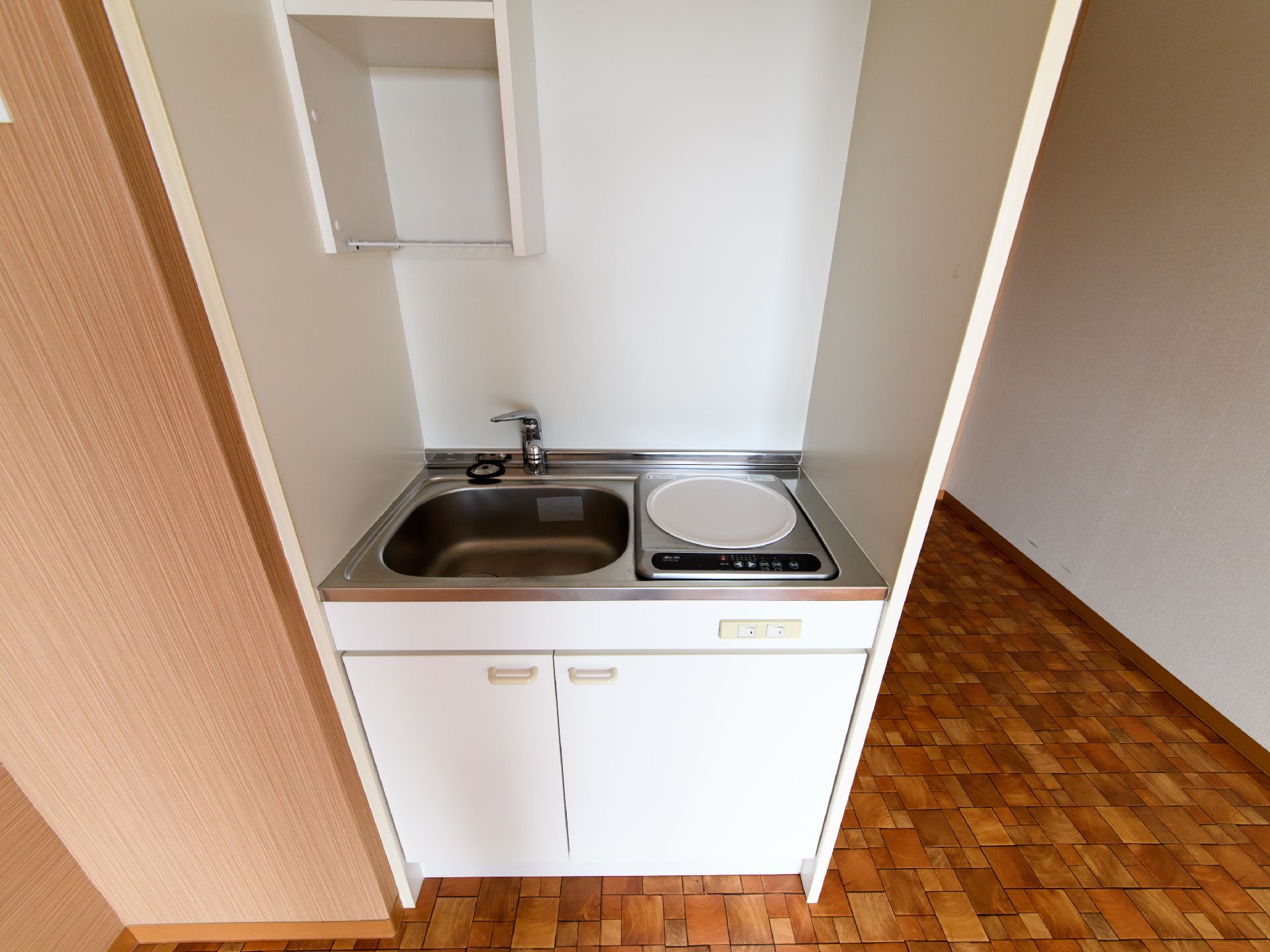 居室内にあるミニキッチン。右側に電気コンロ1箇所、左側に流し台がある。下には開き戸の収納スペースがあり、左上に棚がある。