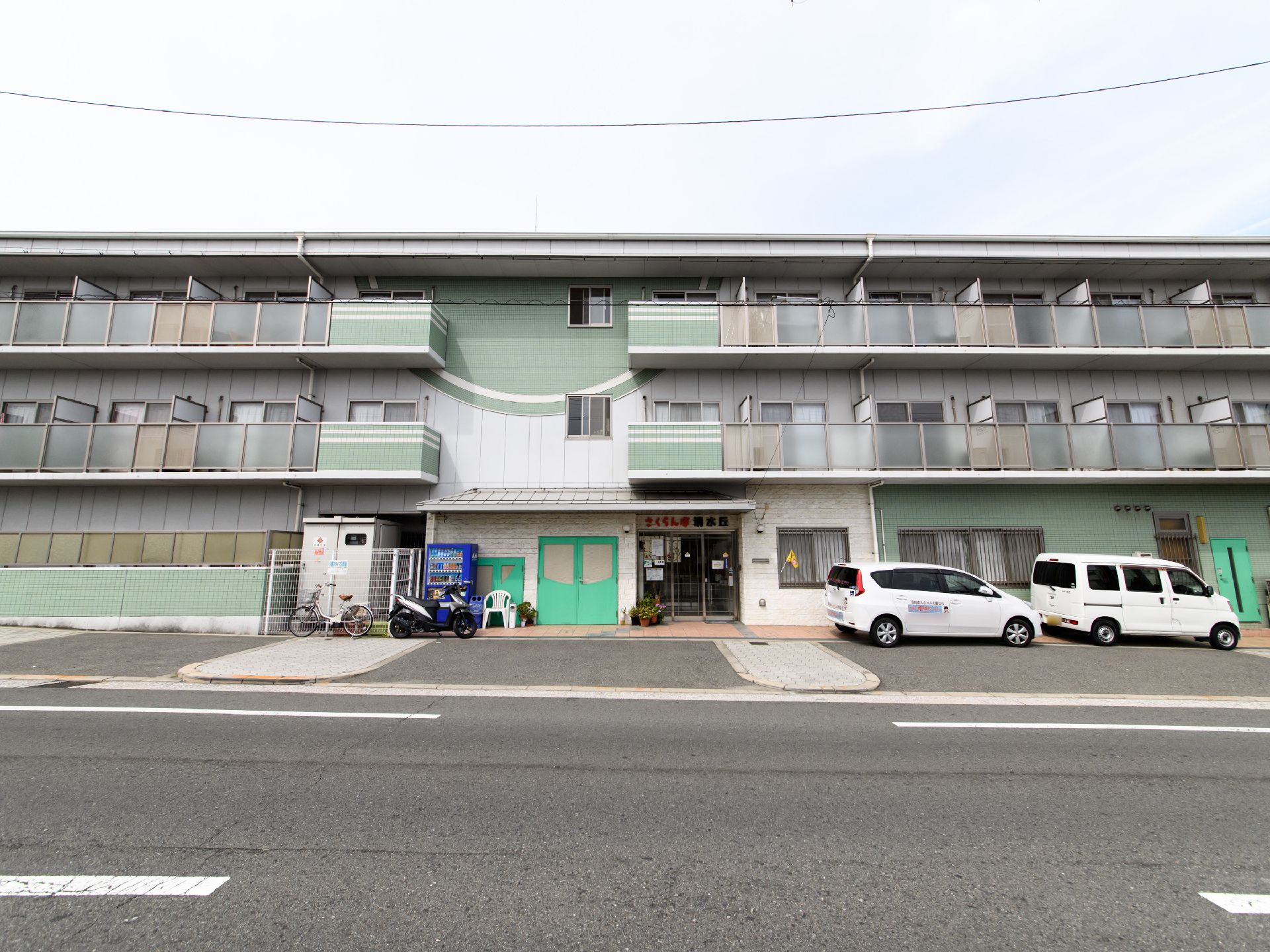 施設は3階建てになっていて、全体は薄い緑色の外観になっている。敷地内には白い車が2台停められている。