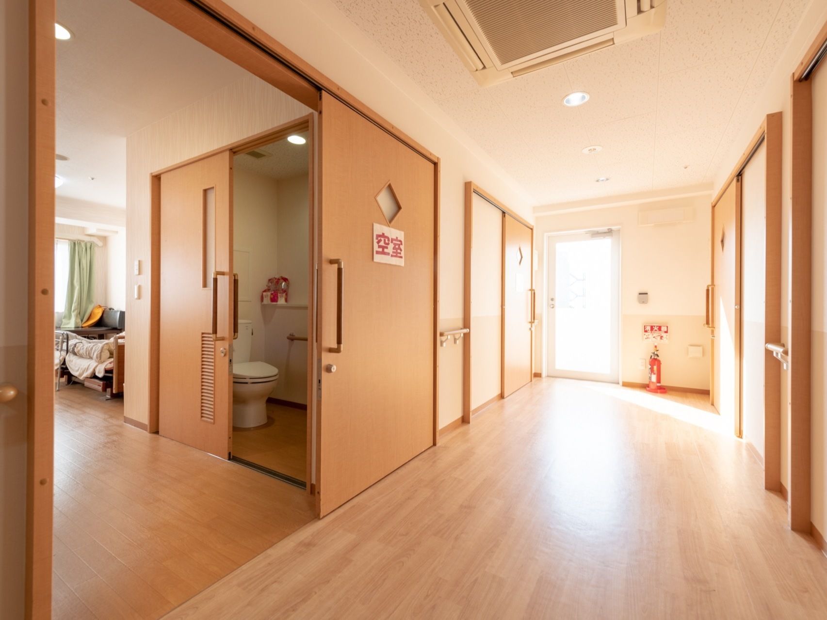 明るい日差しの入る広々とした通路の両側に、居室につながる大きな木目調の引き戸が並んでいる。ドアの上部に小窓がついている。