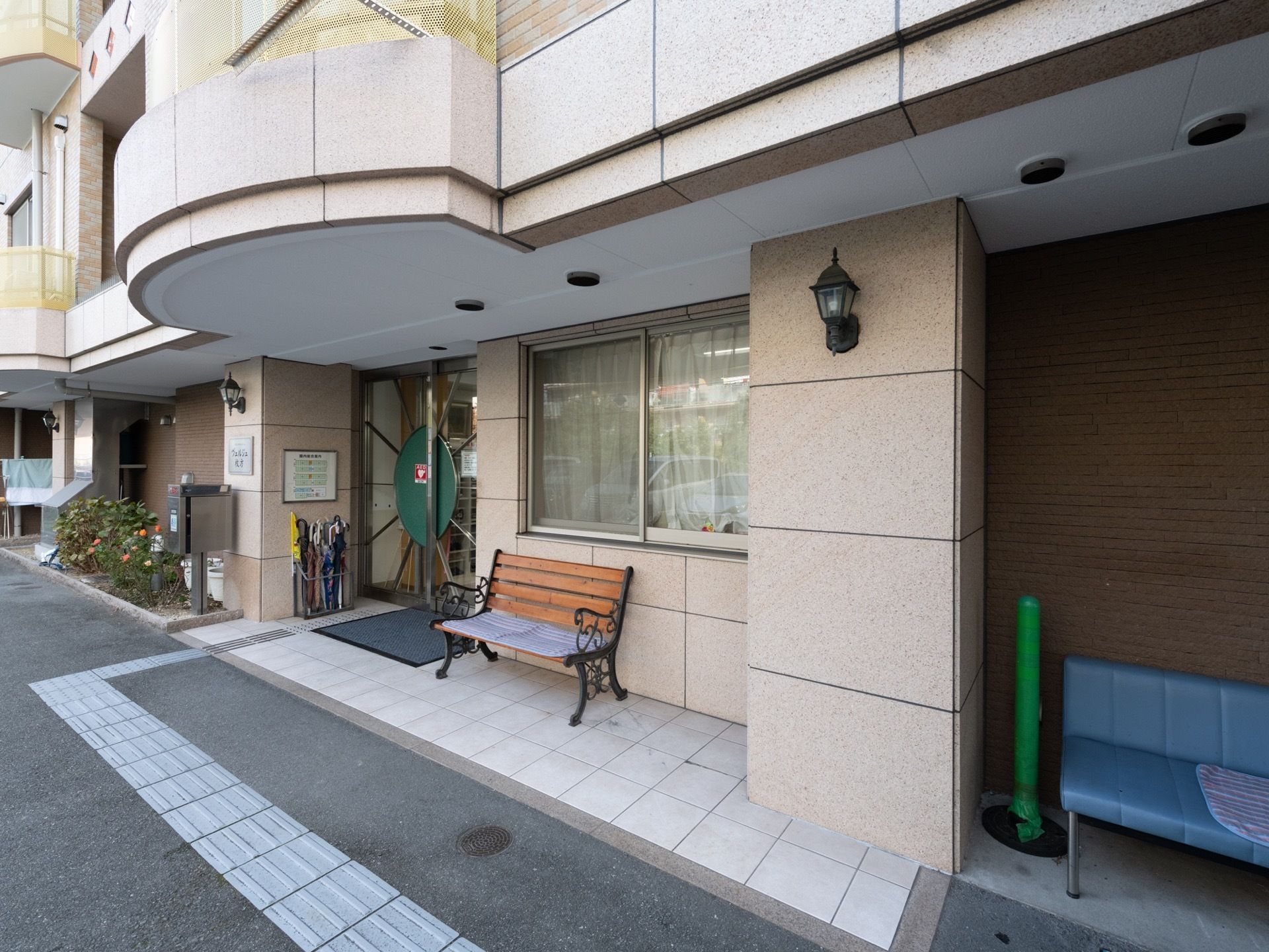 白とベージュの外壁である。玄関のそばには、木製のベンチと傘立て、足元には点字ブロックが設置されている。