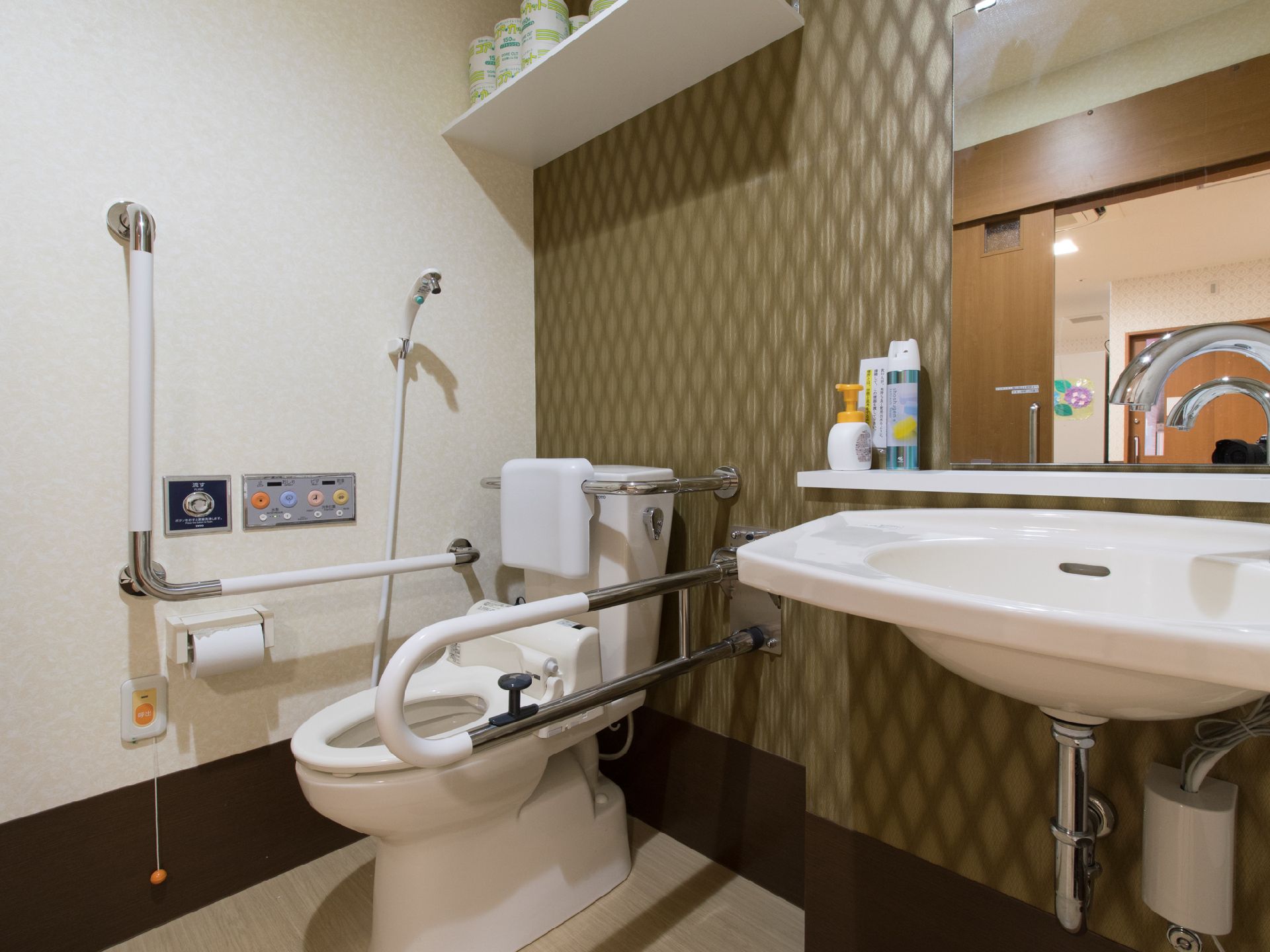 独立した洗面台があるタイプの洋式トイレになっている。手すりは左右にあるので、立ち居が楽である。緊急時のブザーがある。模様がついた壁紙である。