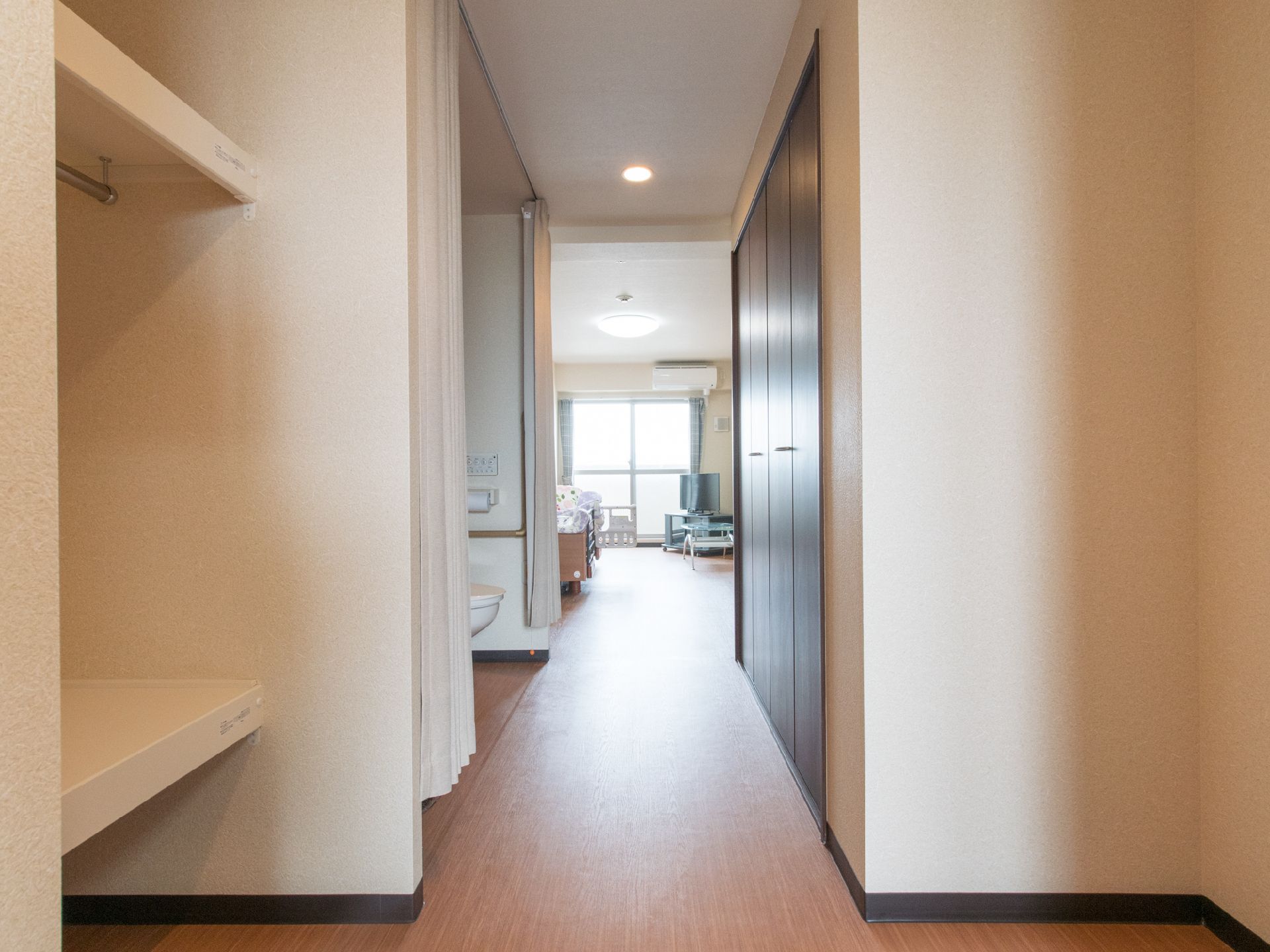 居室で、入り口入ってすぐにトイレ、そのあとに部屋が続いている。壁一枚隔ててトイレとベッドがあり、カーテンで仕切ってある。