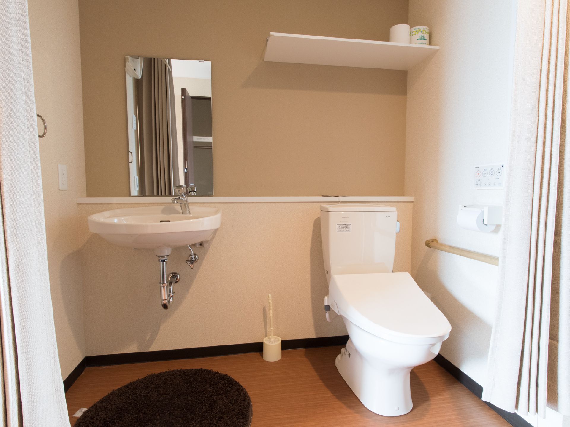 茶色のフロアと白い壁の部屋で洋式トイレがある。隣に大面鏡がついた洗面台が設置してある。壁に手すりがついている。