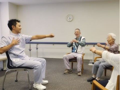 指導員と数名の入居者様が椅子に腰かけて、軽い運動を行っている様子
