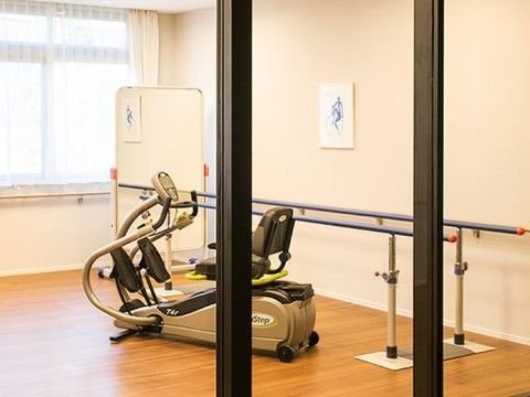 窓にはカーテンがかけられ、壁に手すりが設置されている。室内には2種類のリハビリ用器具と大きな鏡が置かれている。
