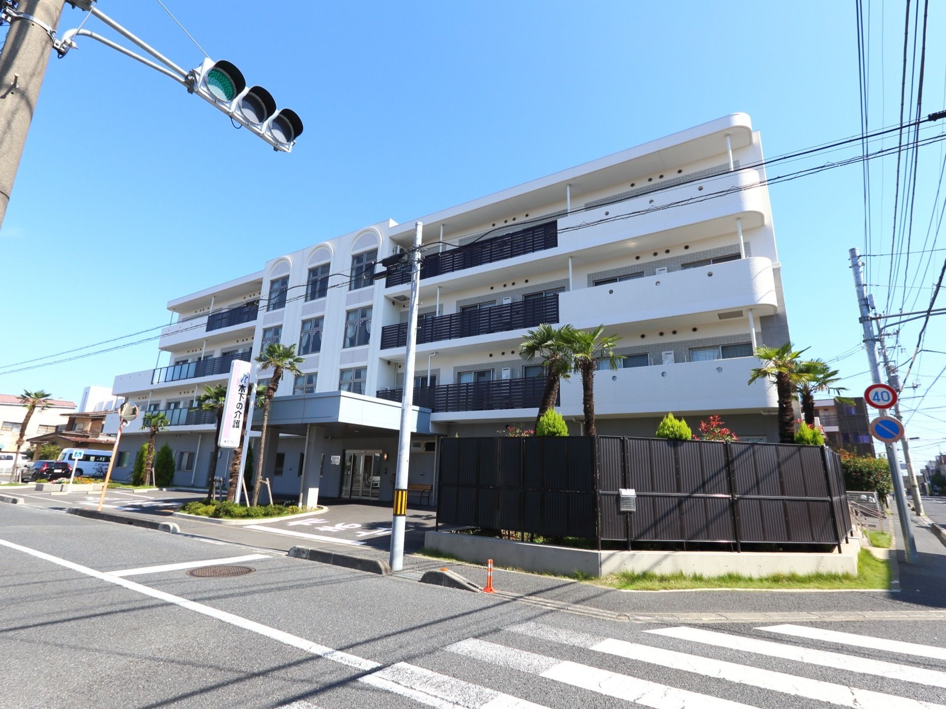 道路に面した白色の外観の４階建ての施設である。敷地内は外部と黒い柵で仕切り、その傍の内側には、植栽を施しており緑豊である。