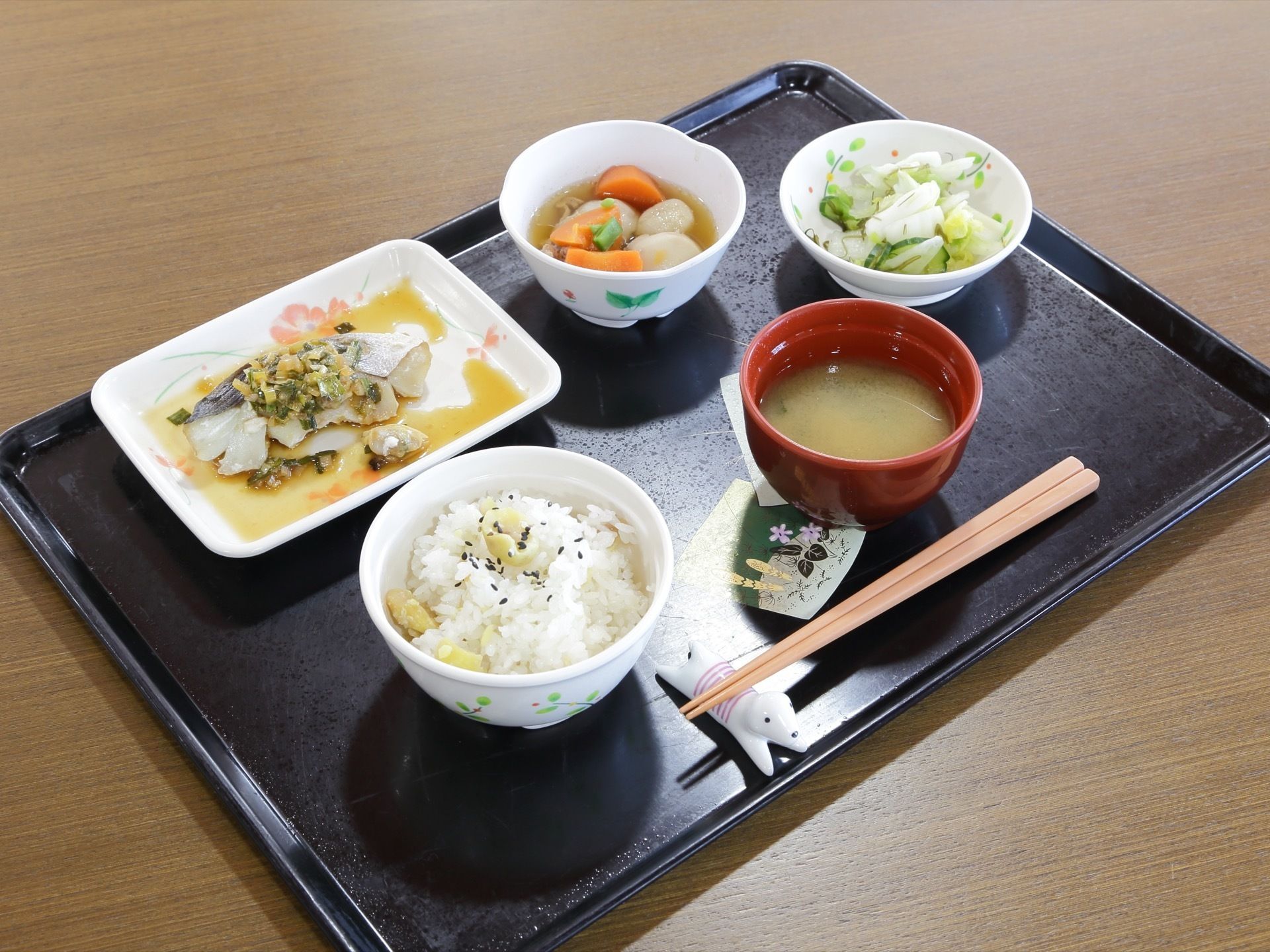 つくしや花などの絵が入った黒色のトレイの上に栗ご飯と味噌汁、魚料理に２つの小鉢、そして、手前に犬の箸置きにお箸をのせて並べている。