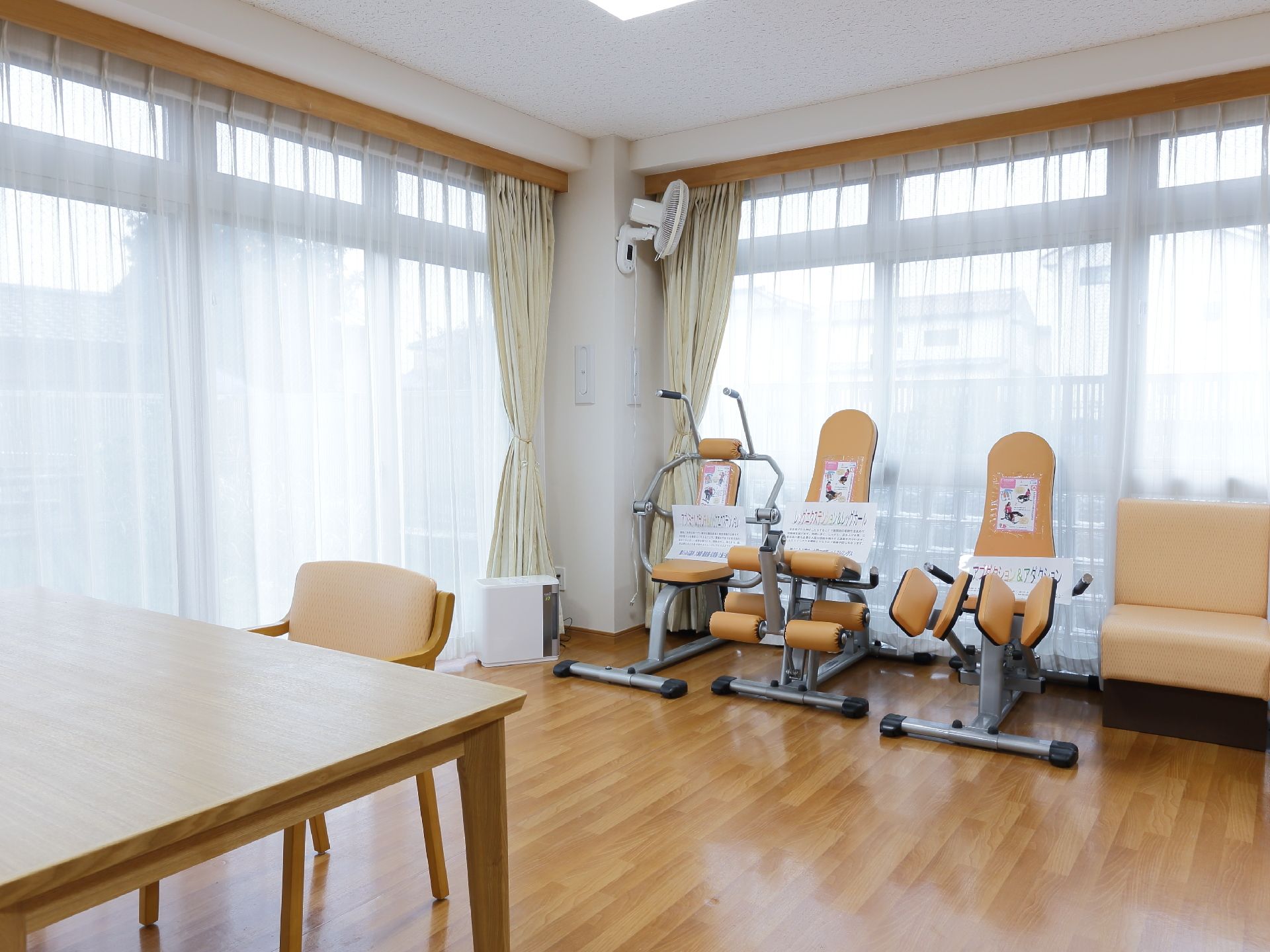 明るい角部屋に位置している共有スペースにはテーブルや椅子が置かれている他、機能訓練機器が３台設置されている。