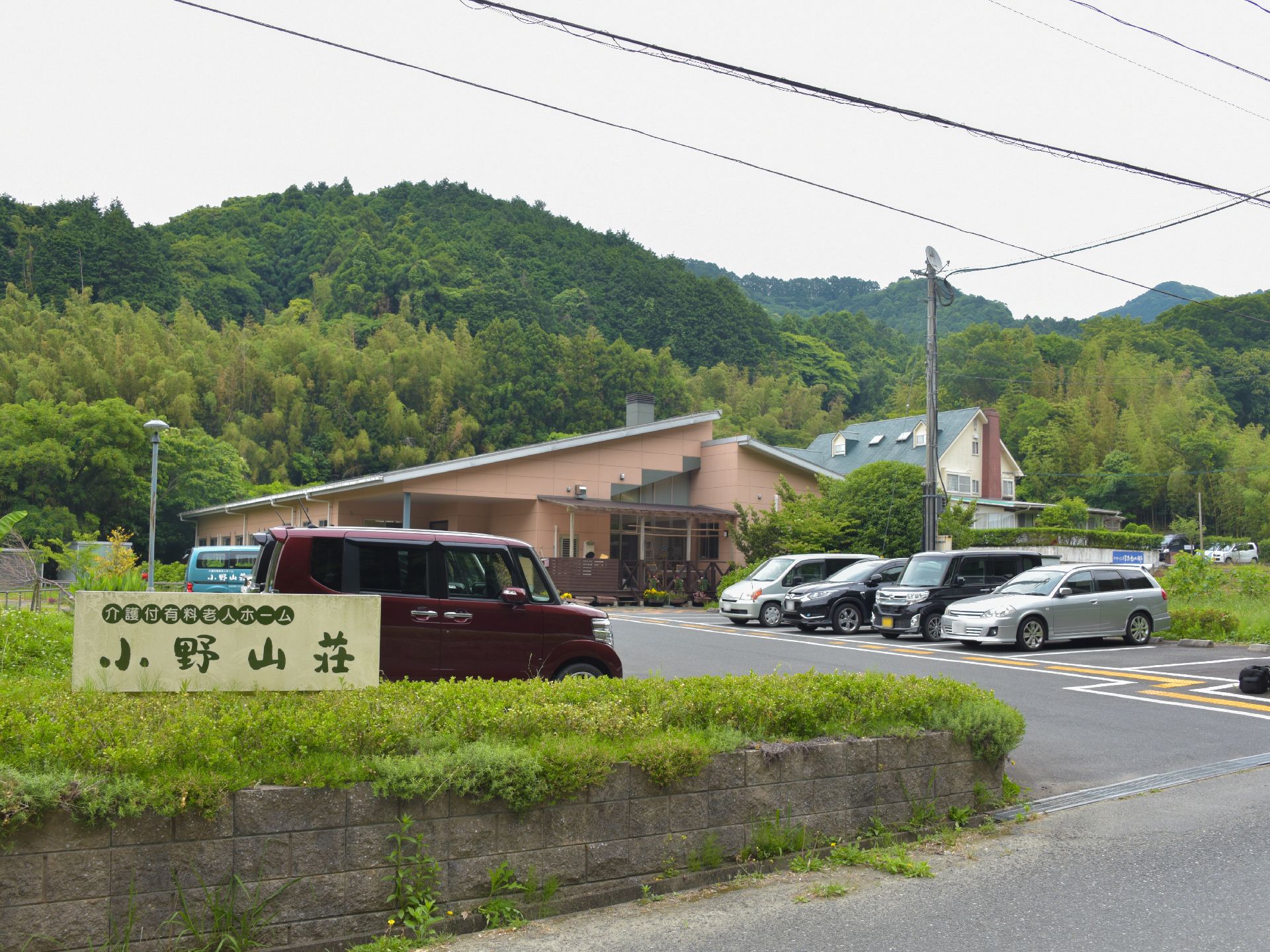 ブロック塀で囲まれたフェンスには施設の名前が掲げてある。建物の前には、駐車場が整備されている。外壁が茶で、緑の屋根が見える。