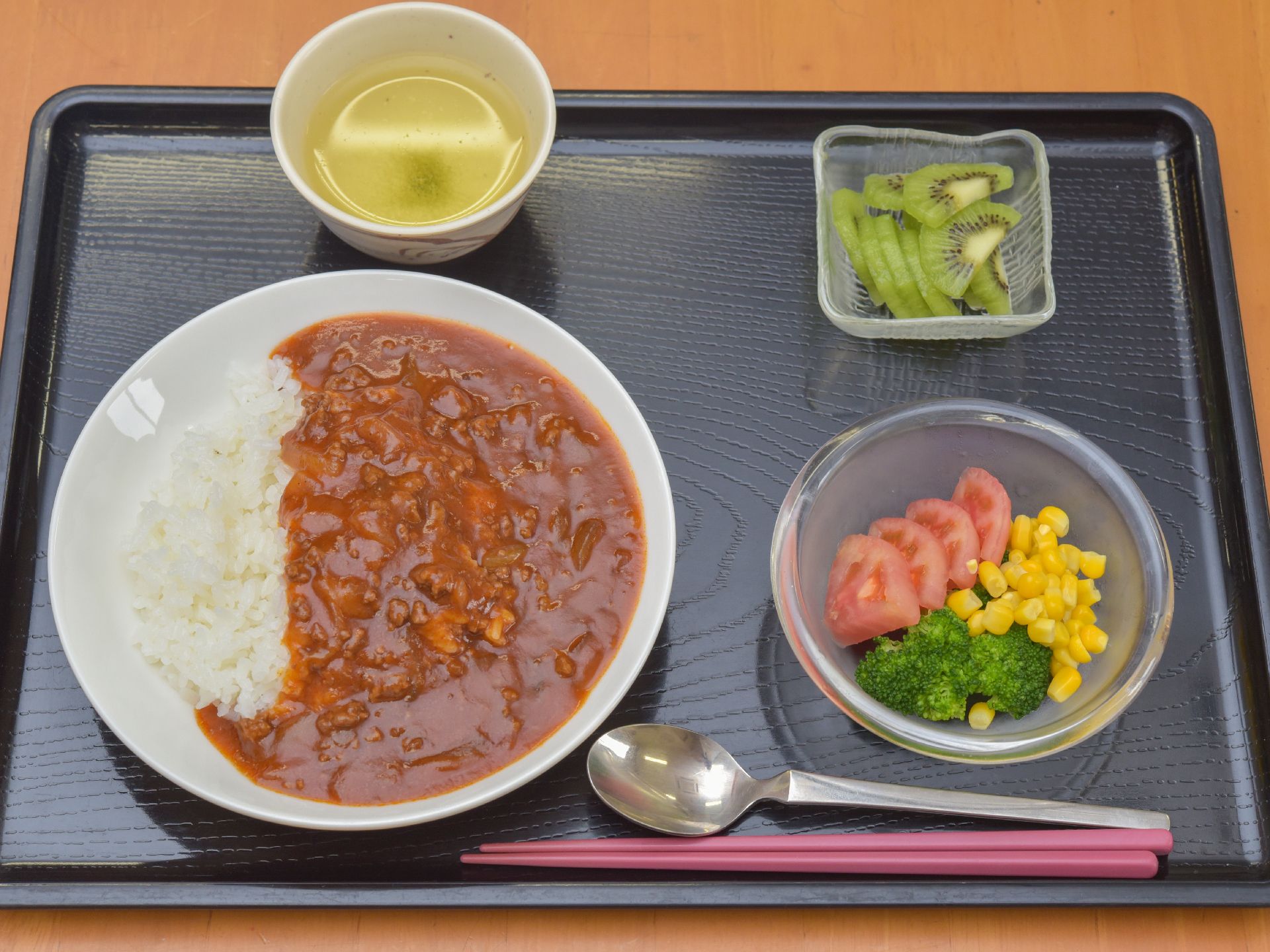 お盆の上にもりつけてある施設の食事の一例で、メインはカレーライスである。湯飲みが置いてある。サラダがついていて、箸とスプーンが置いてある。