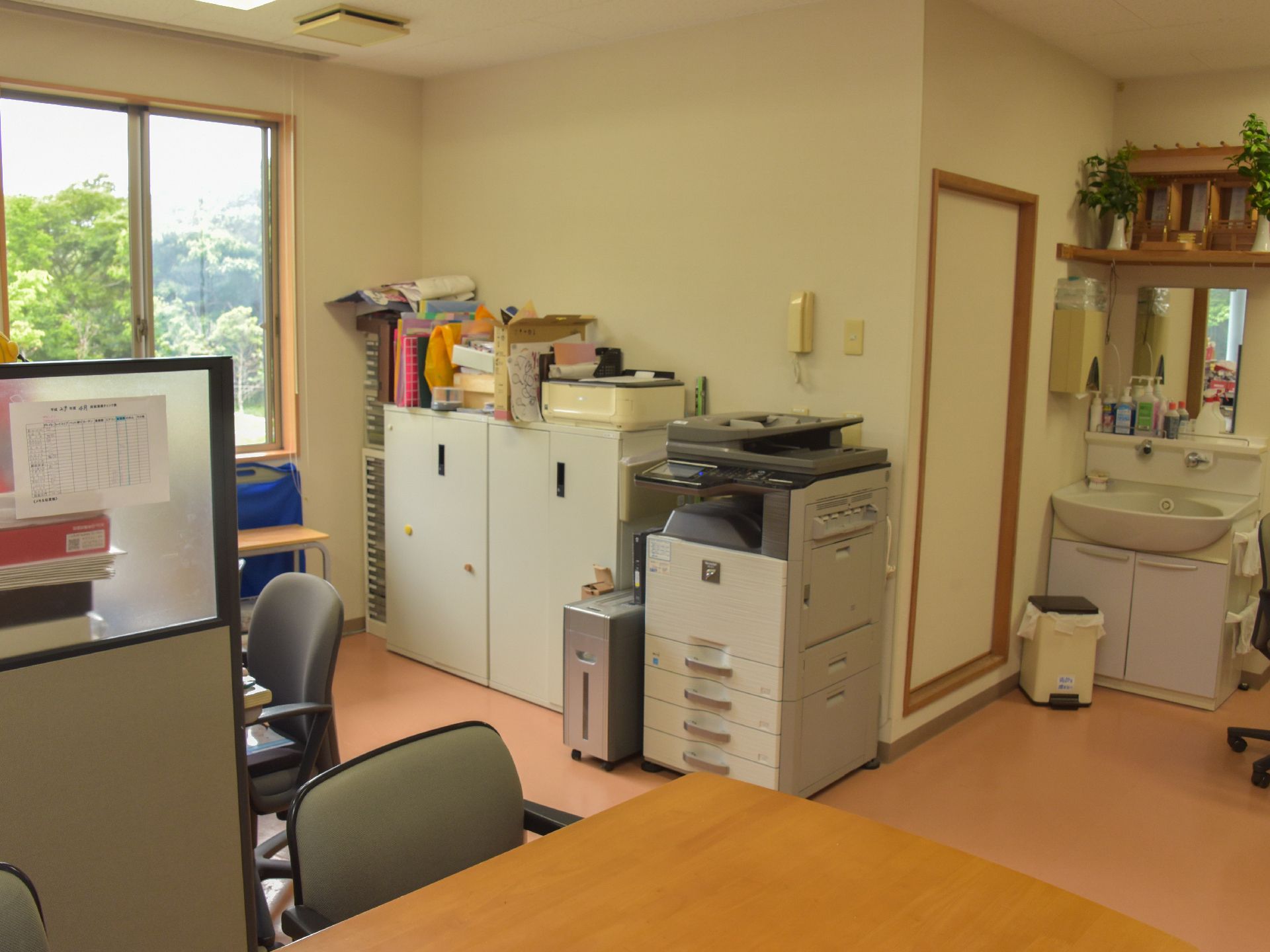 デスクがあり、隣との間は衝立のようなもので仕切ってある。窓からは周囲の緑が見える。コピー機、ファイルキャビネットがなど壁面に置いてある。