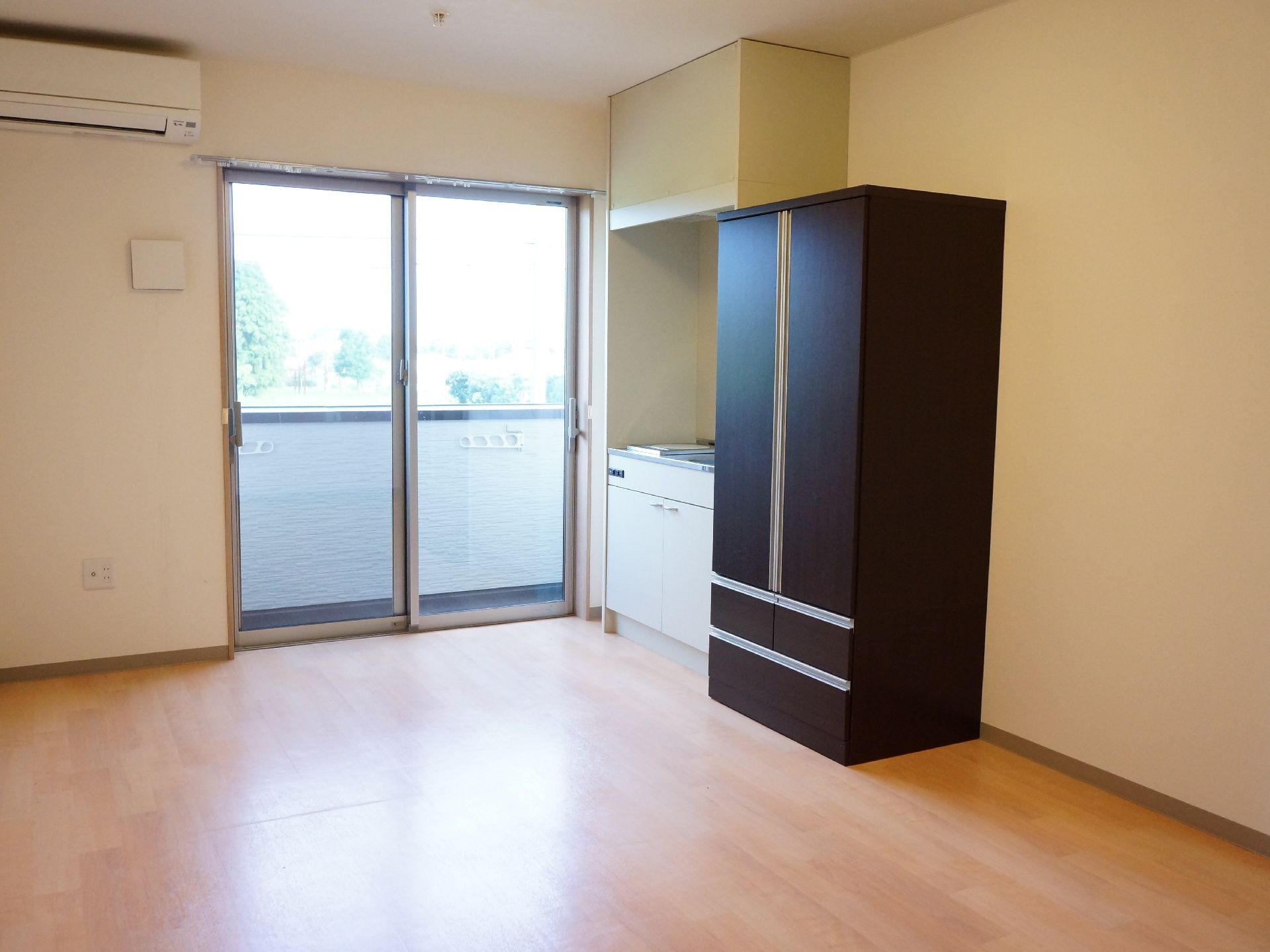 居室はワンルームタイプ。大きな掃き出し窓の外にはベランダがある。壁の上部にはエアコンが設置されている。一角にキッチンと収納がある。