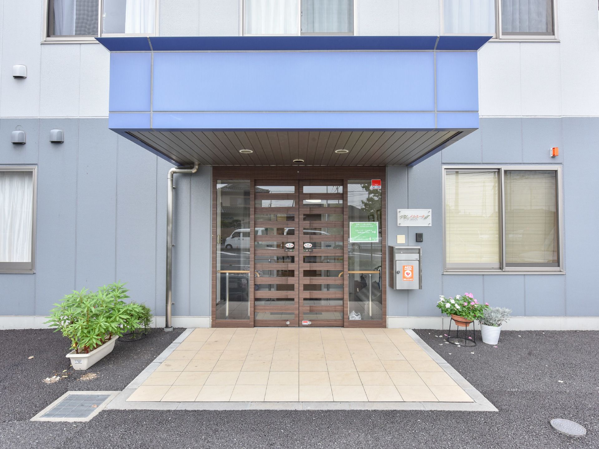 施設の正面には、自動ドアの上の部分に青い色をした雨除けの屋根が設置されている。両サイドには観葉植物が置いてある。