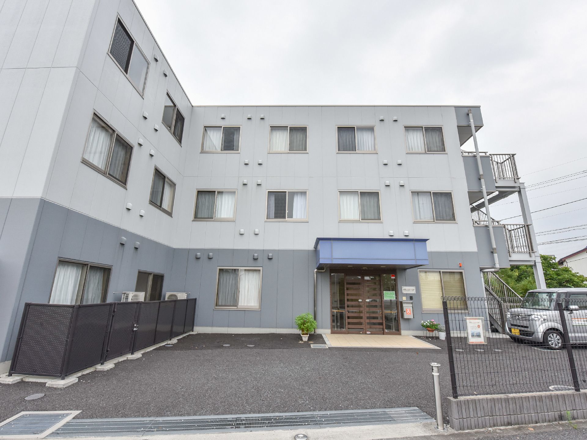 施設は黒いフェンスで囲われている。建物の外観はグレー色で、入り口には自動ドアが設けられている。車がある。