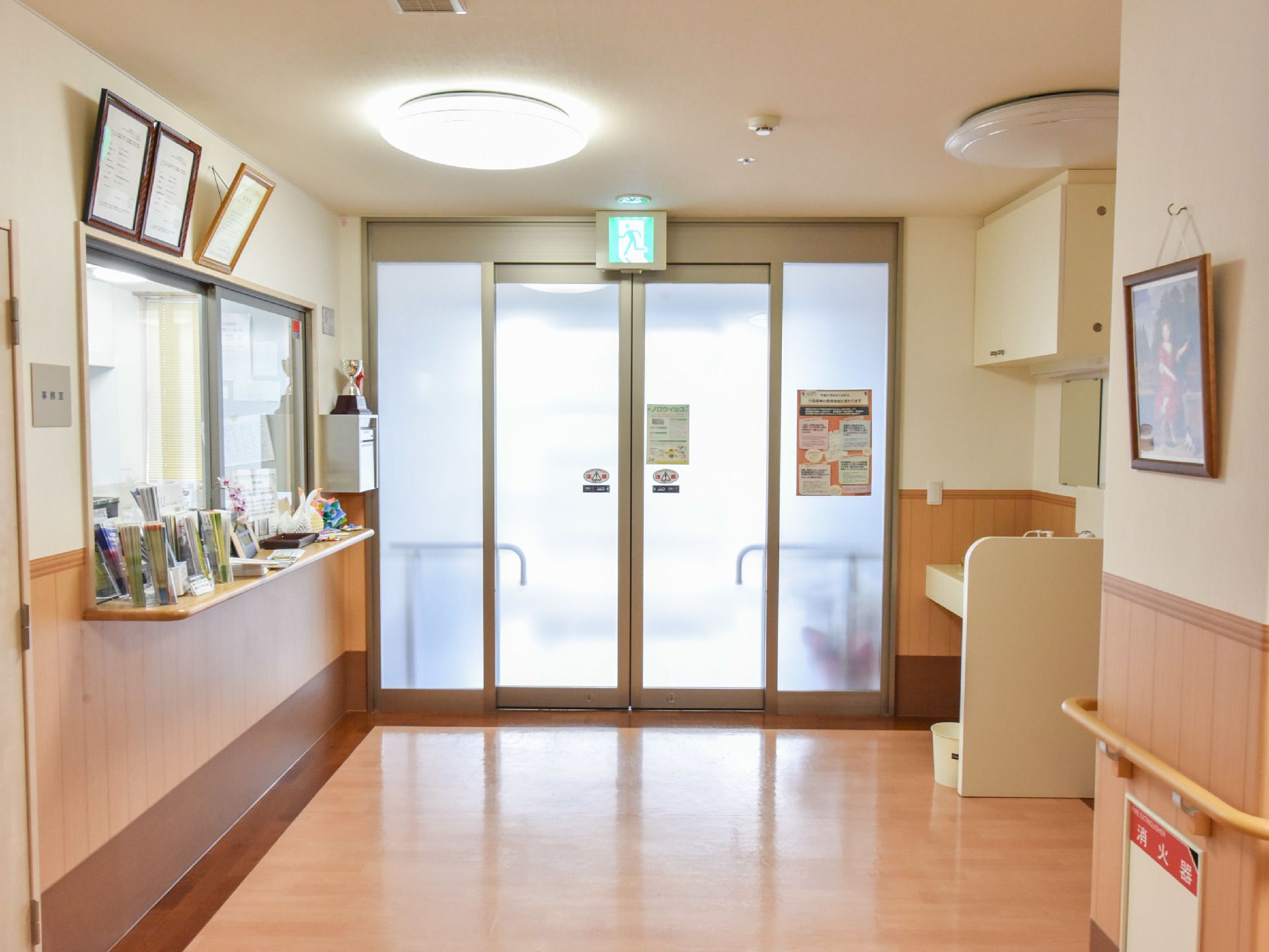 施設入口の正面には自動ドアがあり、非常口の看板も吊り下がっている。右手の壁には絵画や消火器、左手には事務室がある。