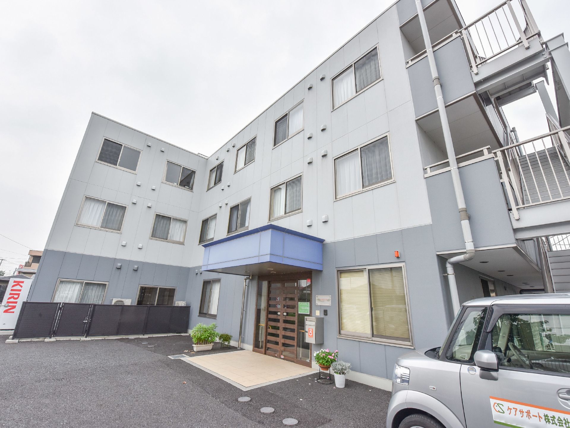 施設を横側から見た形では、3階建てで螺旋型の非常階段も見える。右下には施設専用の車が駐車されている。