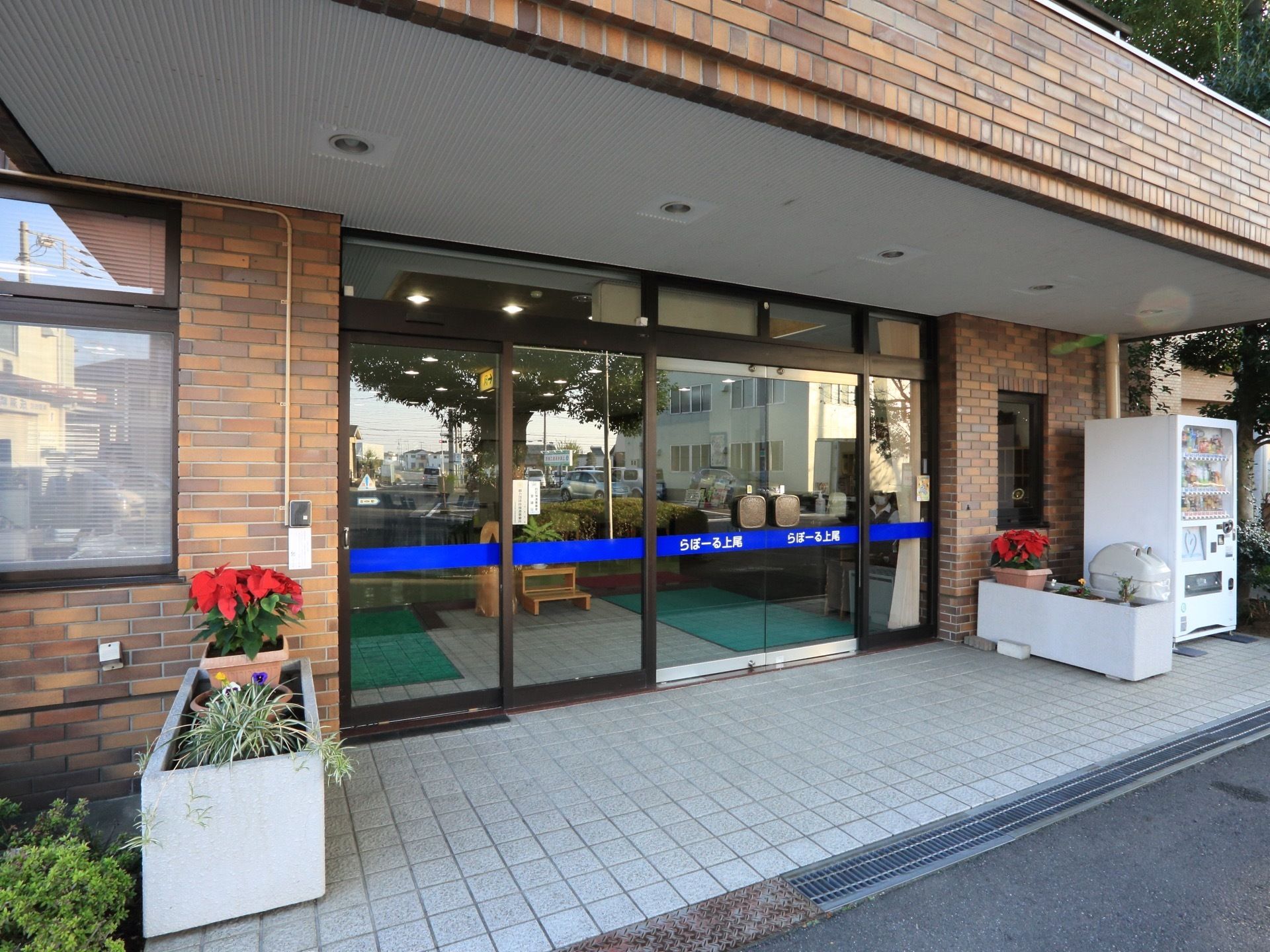 玄関の扉はガラス張りになっており、施設名をわかりやすく記載している。扉の両脇に赤い花が飾られ、華やかになっている。