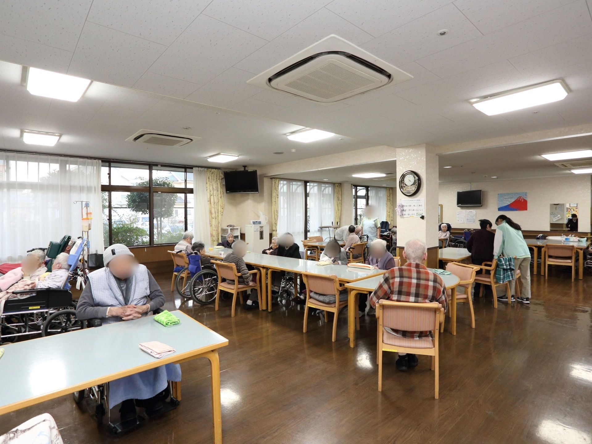 テーブルと椅子が設置してある部屋でたくさんの入居者様がくつろいでいる。スタッフがそのお世話をしている。