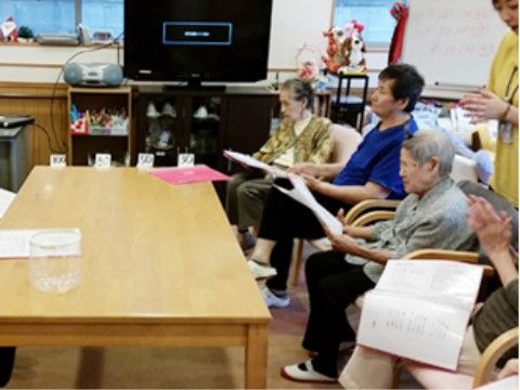 椅子に腰掛け冊子を手にもち、手拍子したり見つめたりしている入居者様