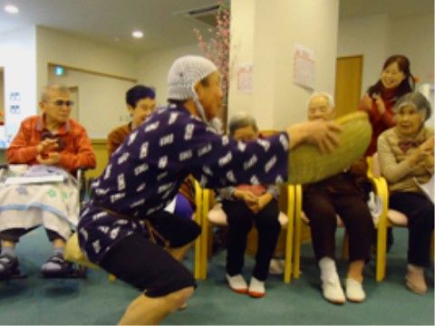 椅子に腰掛け手拍子する入居者様の前で、季節イベントの踊りを披露するボランテイアの男性