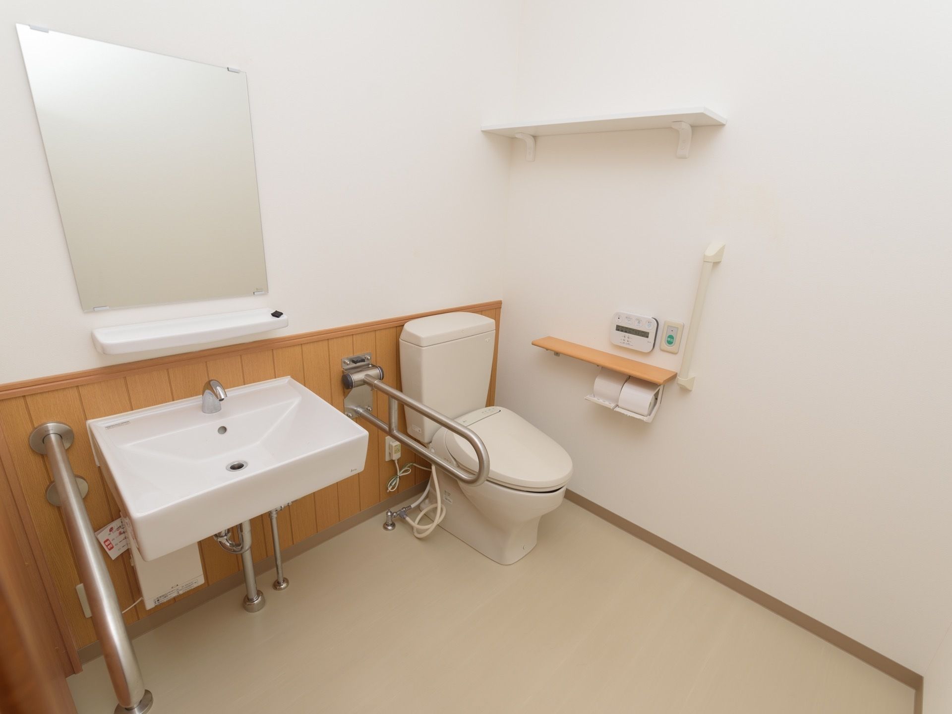 白い壁に囲まれた広いトイレスペースに蓋付きの洋式トイレが設置されている。壁面には、小さな物を置くことができる棚がある。